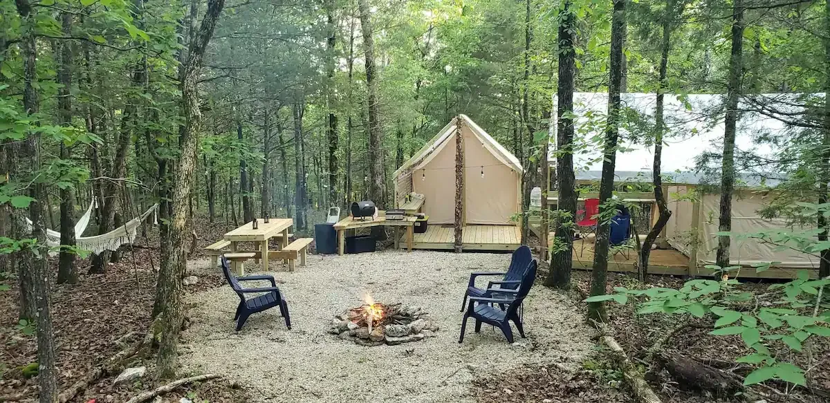 Wooded campsite with a tent, picnic table, fire pit, and two chairs among trees