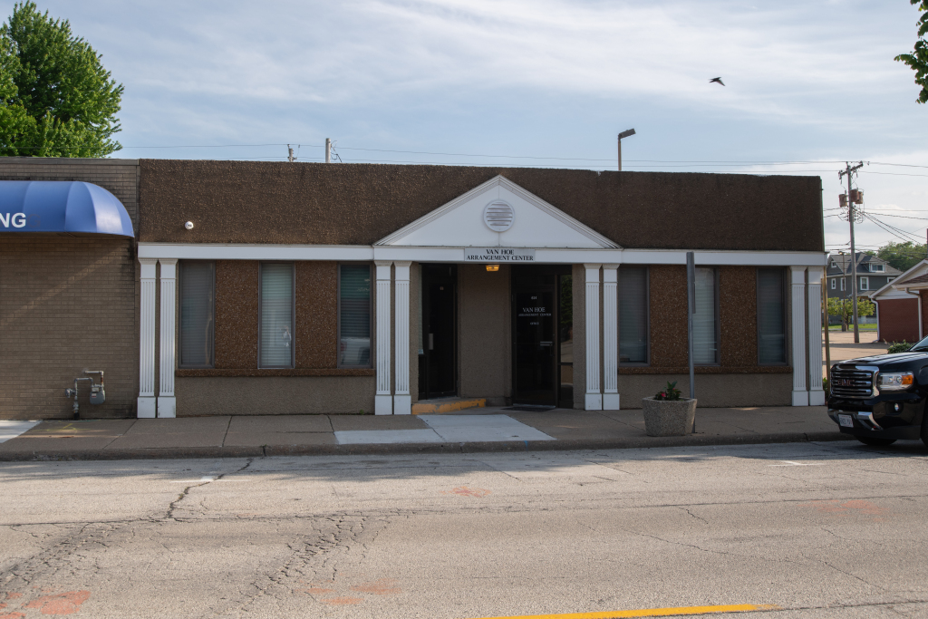 A one-story brick building with a brown roof and white pillars; a police car is parked to the right.