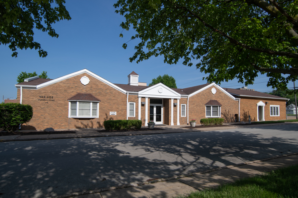 Brick building with multiple gabled sections, white trim, and a prominent entrance under a small cupola, trees casting shadows on the paved area.