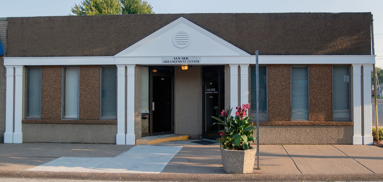 Building with white pillars, brown roof and facade, open doorway and a flower pot.