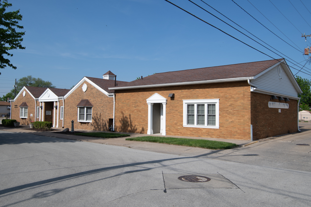 Tan brick building with brown roof and white trim. Blue sky and power lines overhead.