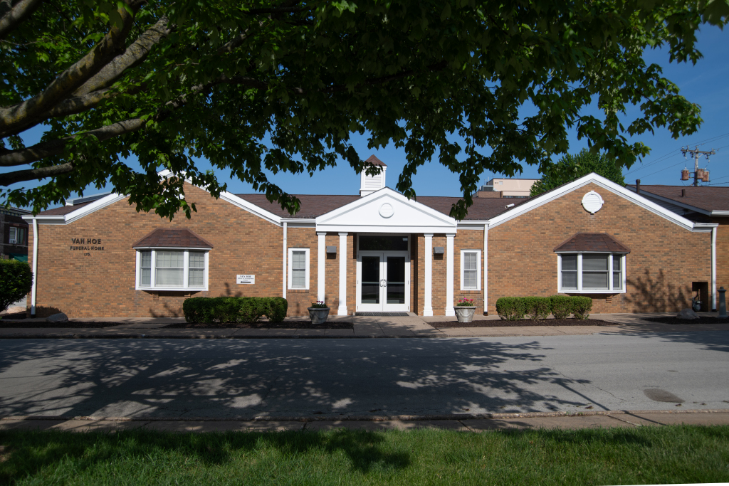 Tan brick building with white trim, arched roof, and double doors. Tree casts shadows.