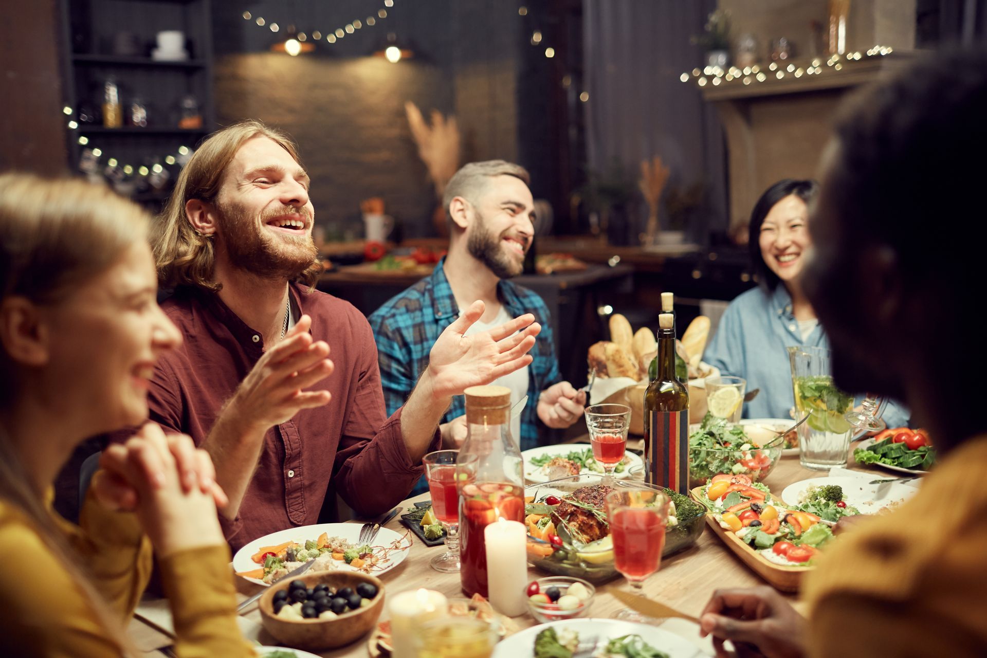 People at a dinner party, laughing, clapping. Table with food, drinks, and candles. String lights in background.