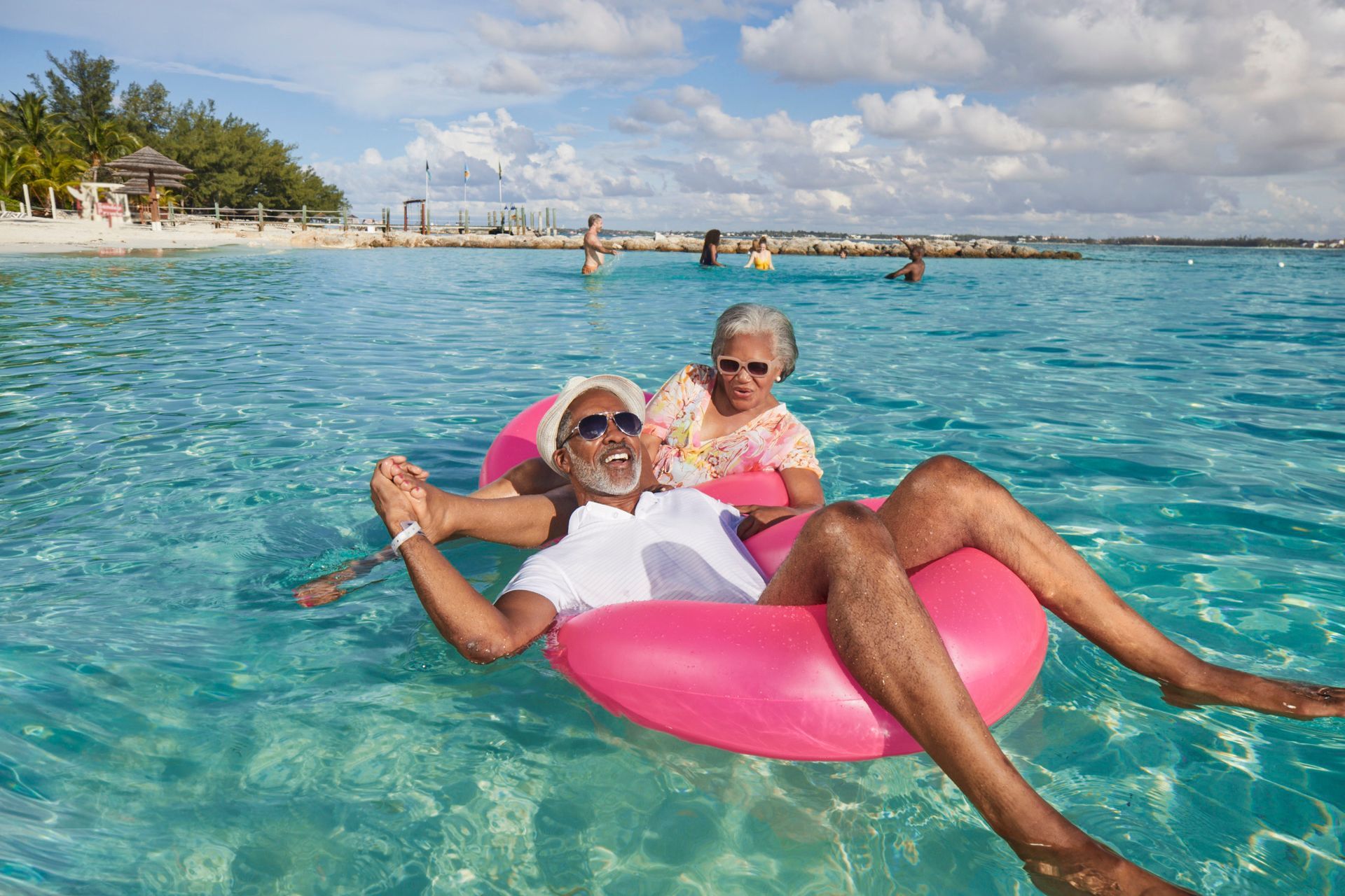 Two people lounging on a pink float in clear turquoise water near a tropical beach