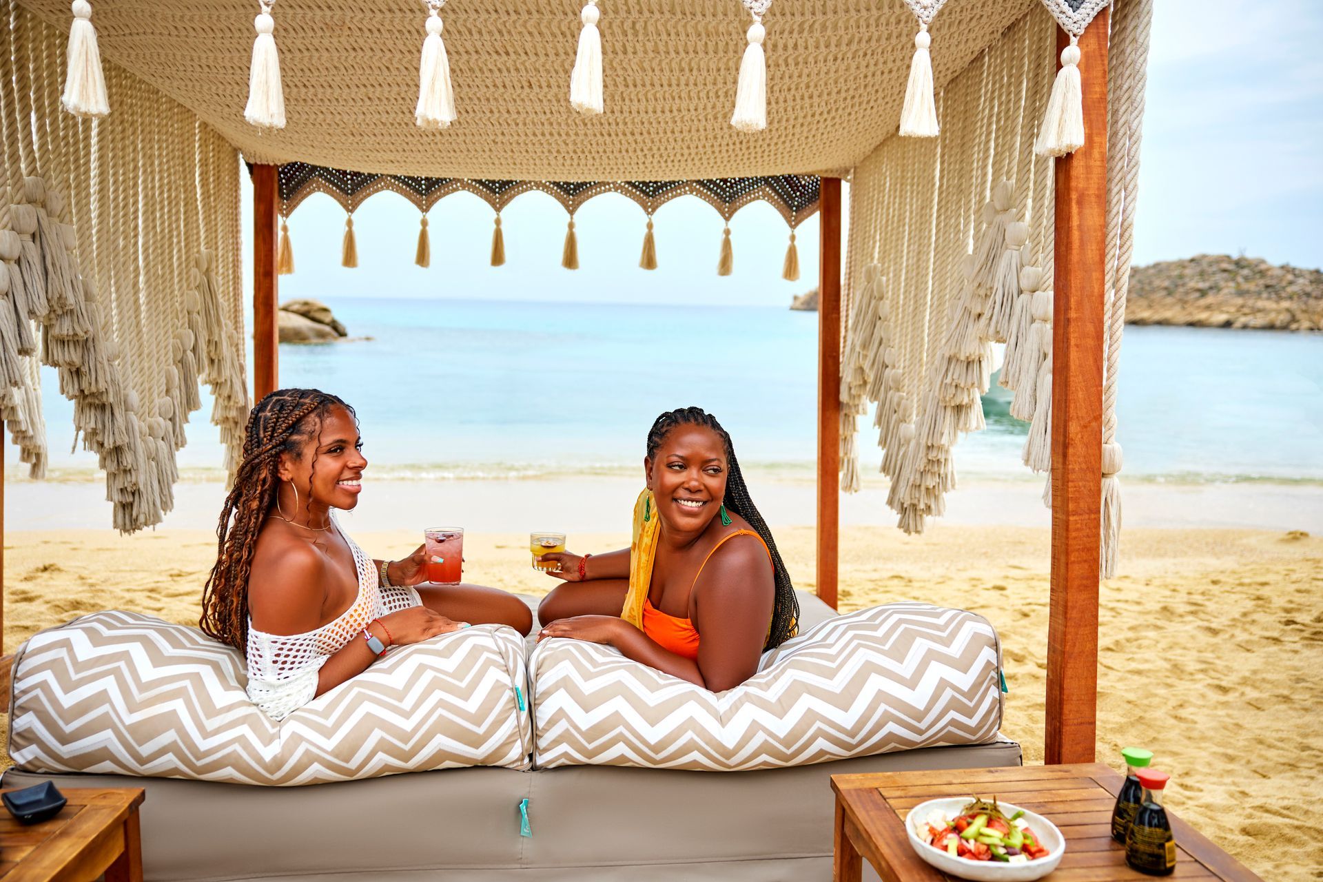 Two people relaxing on a beach daybed under a canopy, with the sea and sandy shore behind them.