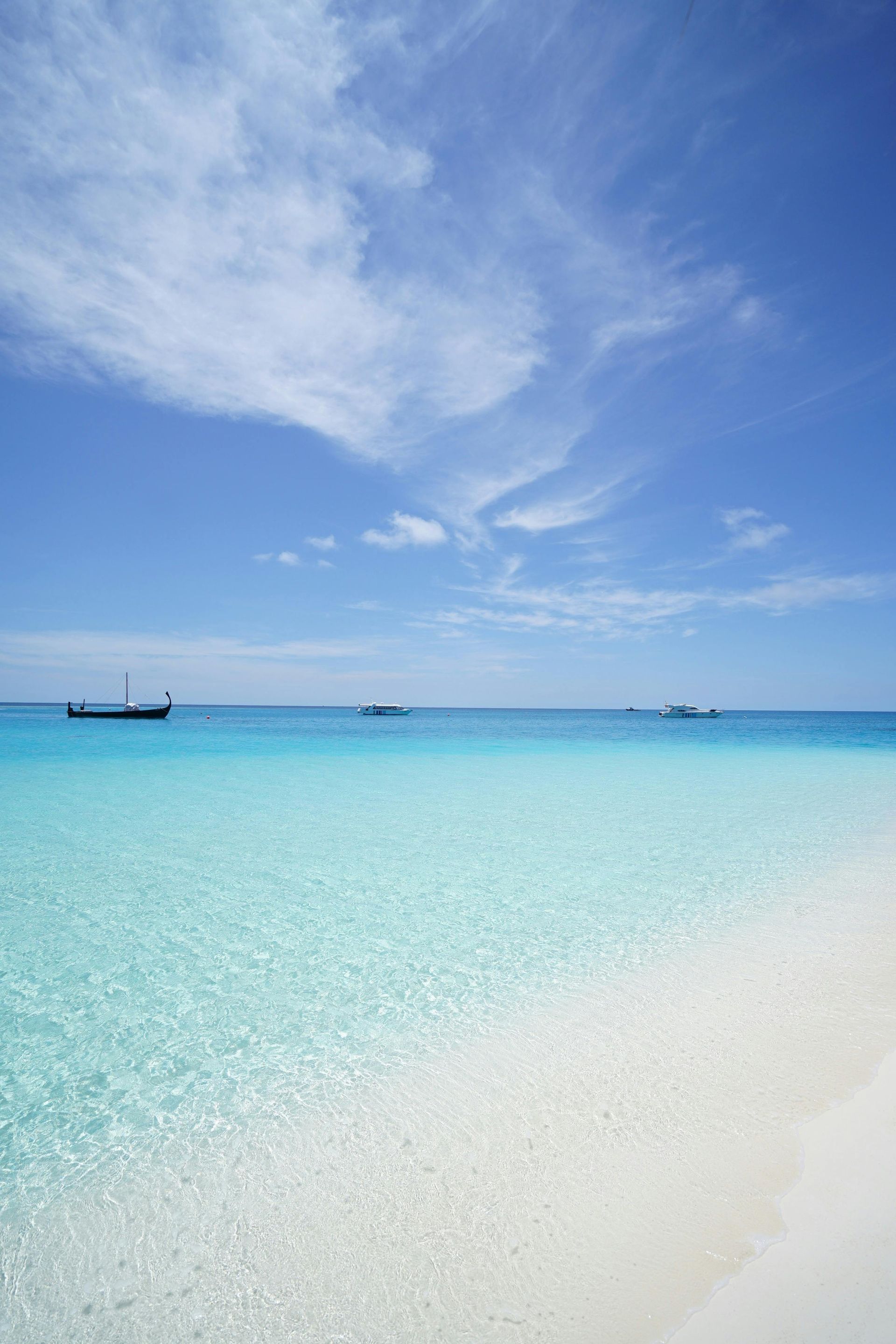 Turquoise ocean meeting white sandy shore under a bright blue sky with wispy clouds and distant boats