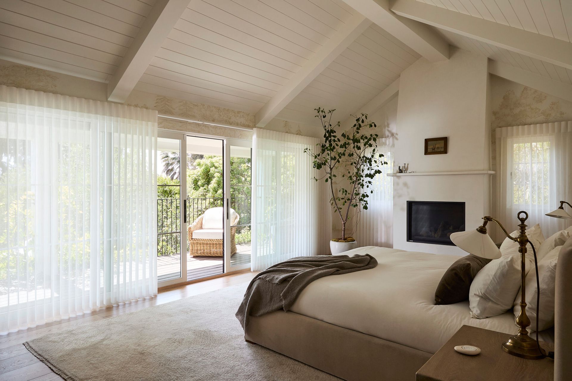 Bright bedroom with vaulted ceiling, sheer curtains, a bed, fireplace, and open doors to a patio.