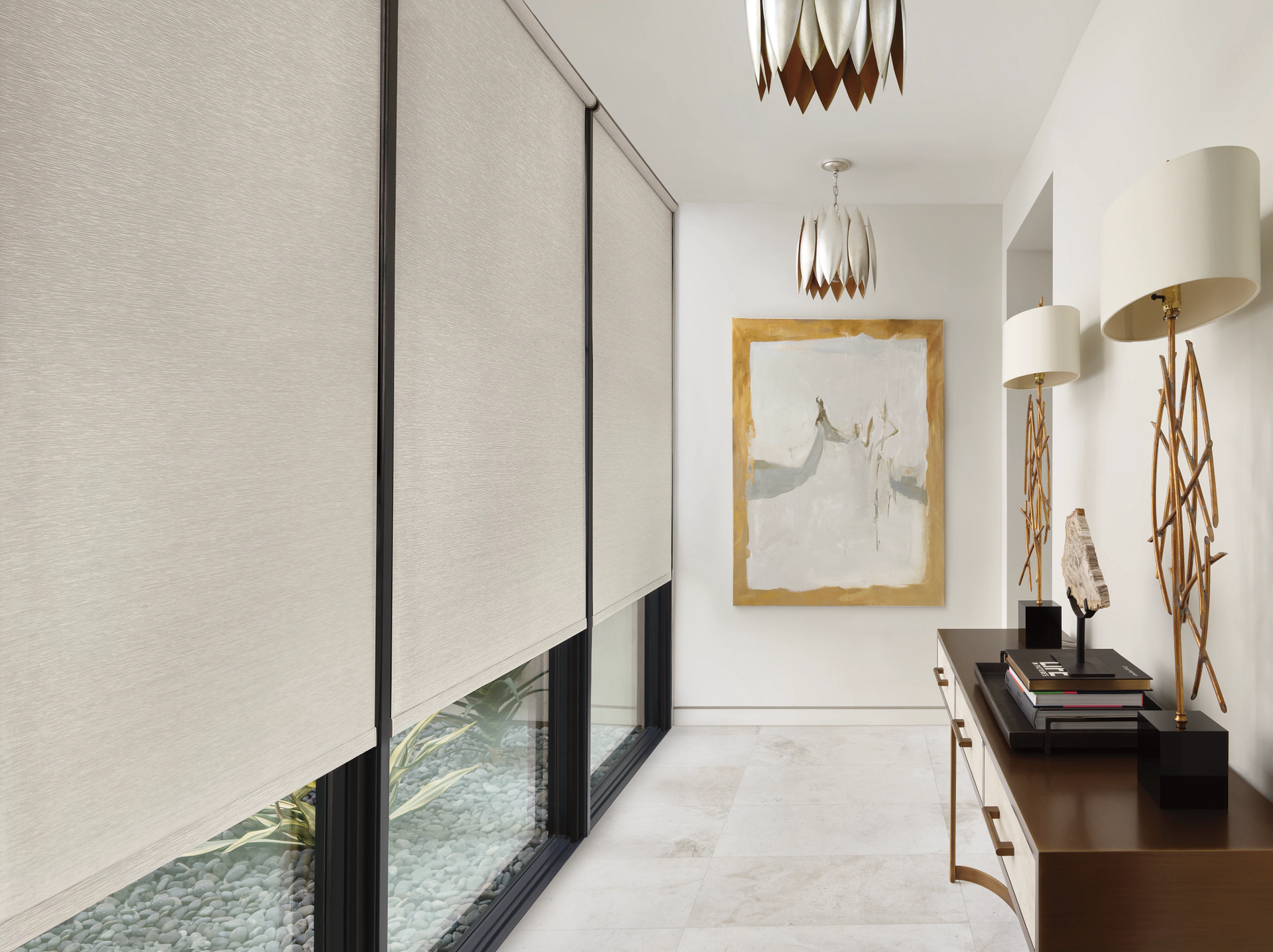 Modern hallway with rolled blinds, marble floor, gold-framed art, and a dark console table with lamps