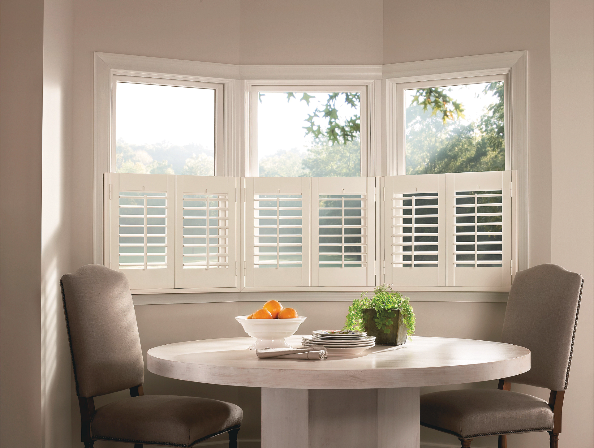 Bright dining nook with bay windows, white shutters, round table, and two beige chairs.