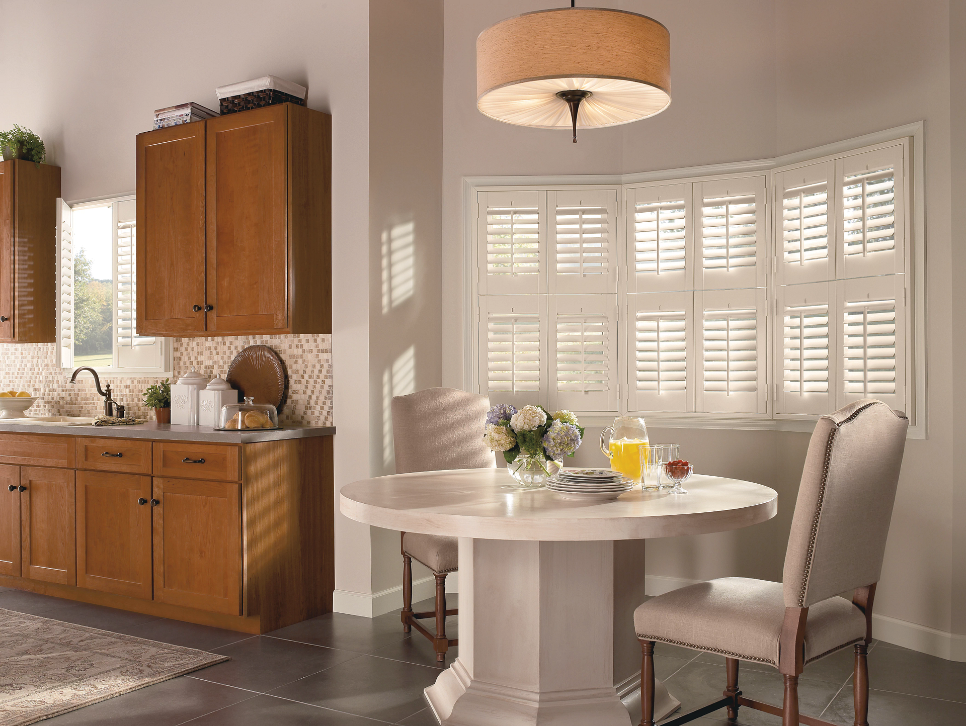 Bright kitchen dining nook with a round table, two chairs, and white plantation shutters in warm sunlight