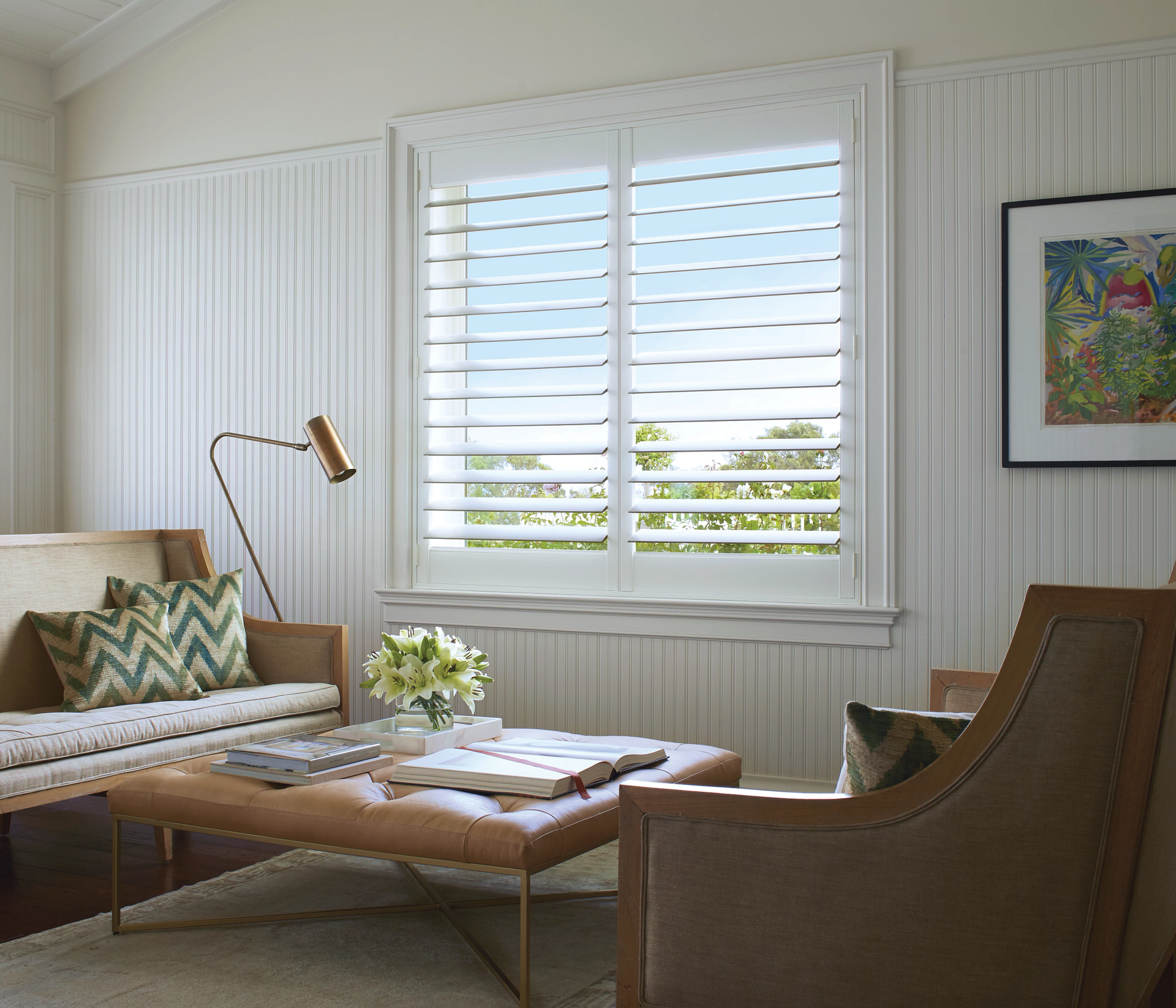 Bright living room with white shutters, gray sofa, coffee table, and wooden chair by a window