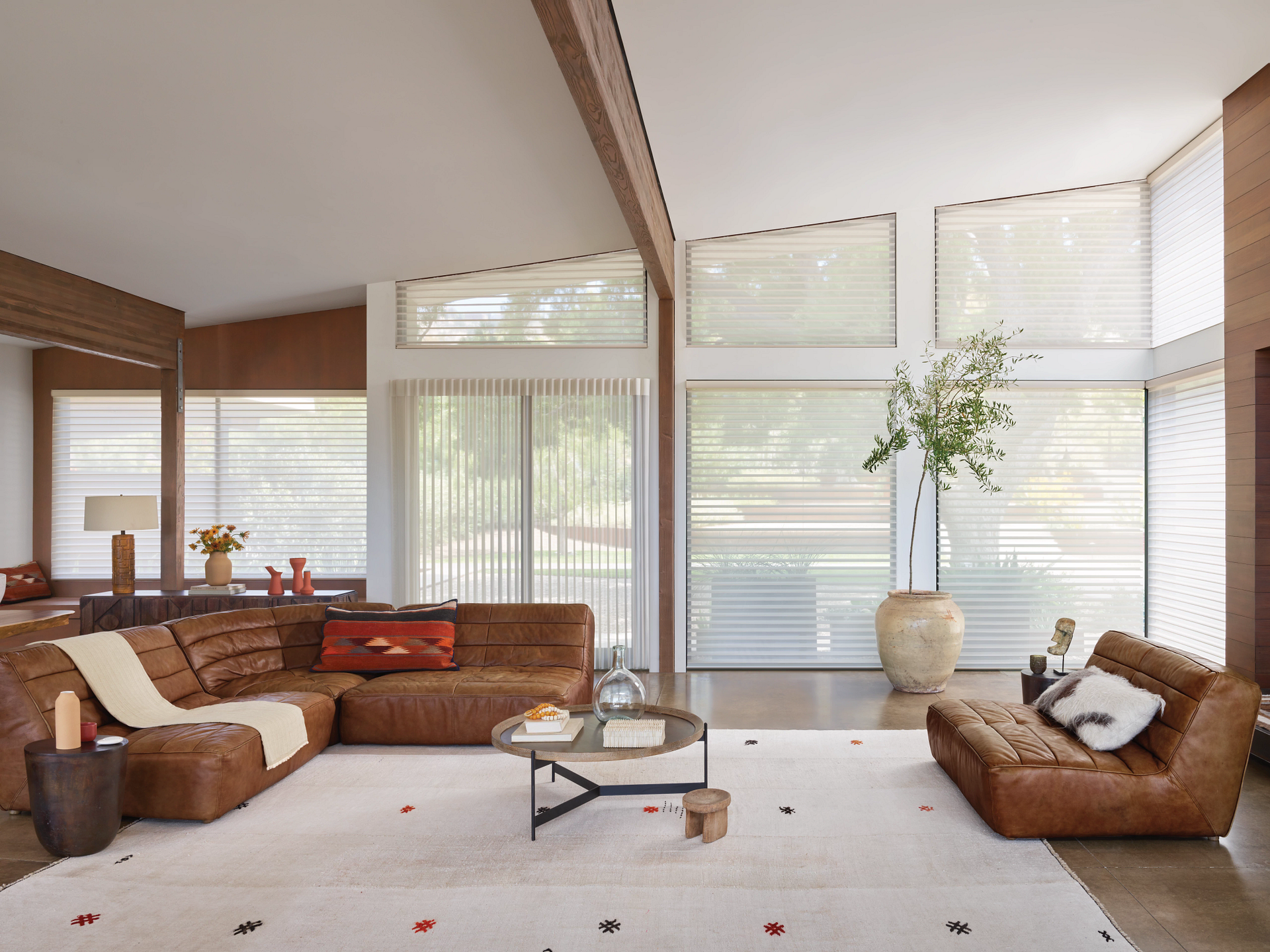 Bright living room with large windows, white blinds, brown sectional, and low leather chairs around a coffee table.
