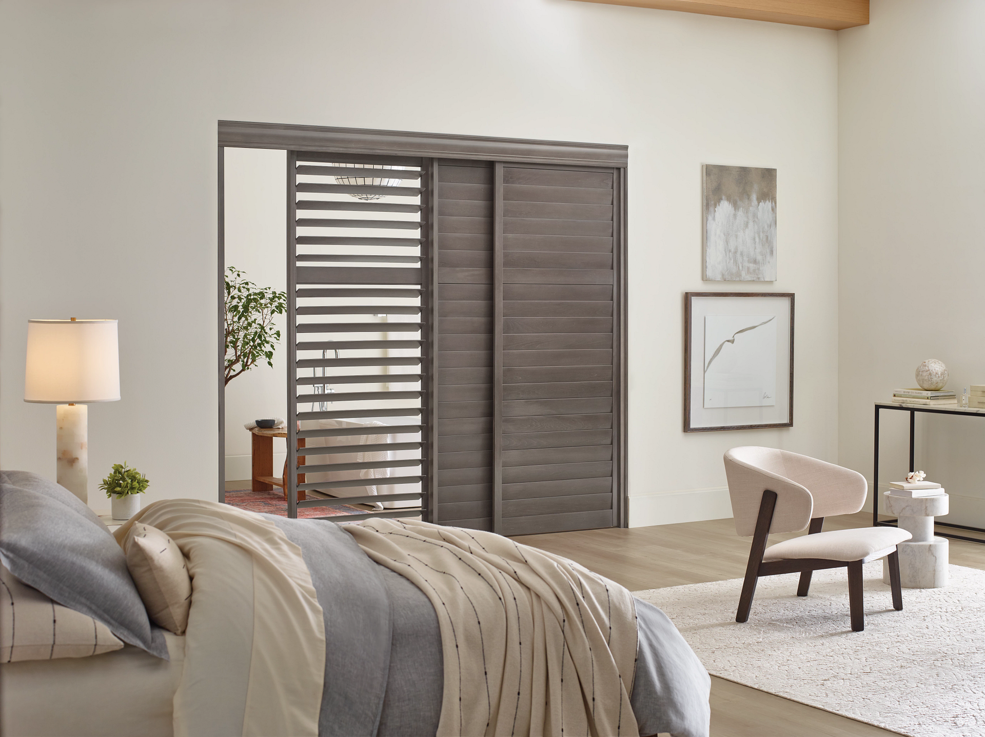 Modern bedroom with beige bedding, wood shutters, lamp, chair, and minimalist wall art