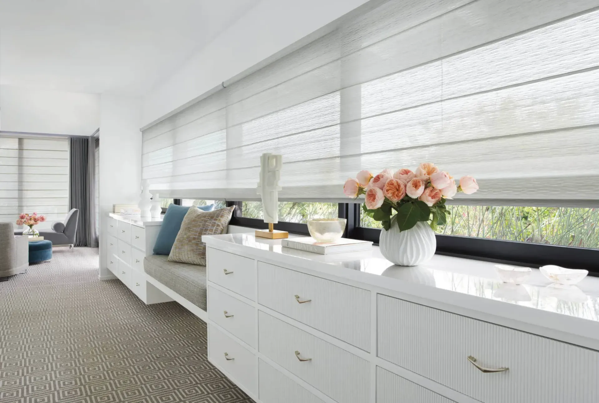 Bright white bedroom with built-in bench, dresser, and large windows with blinds and flowers