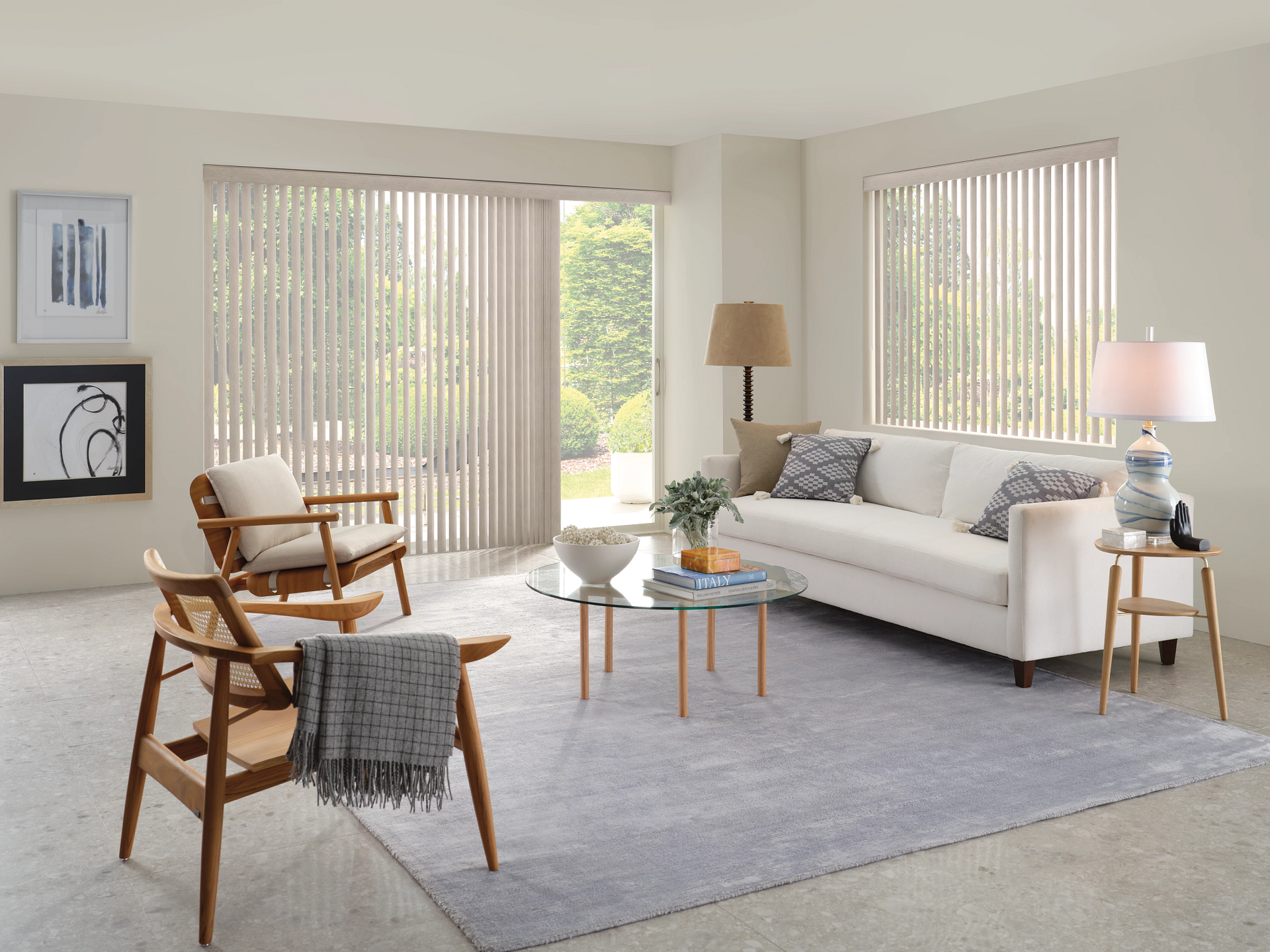 Bright living room with white sofa, wooden chairs, gray rug, side tables, and vertical blinds by tall windows