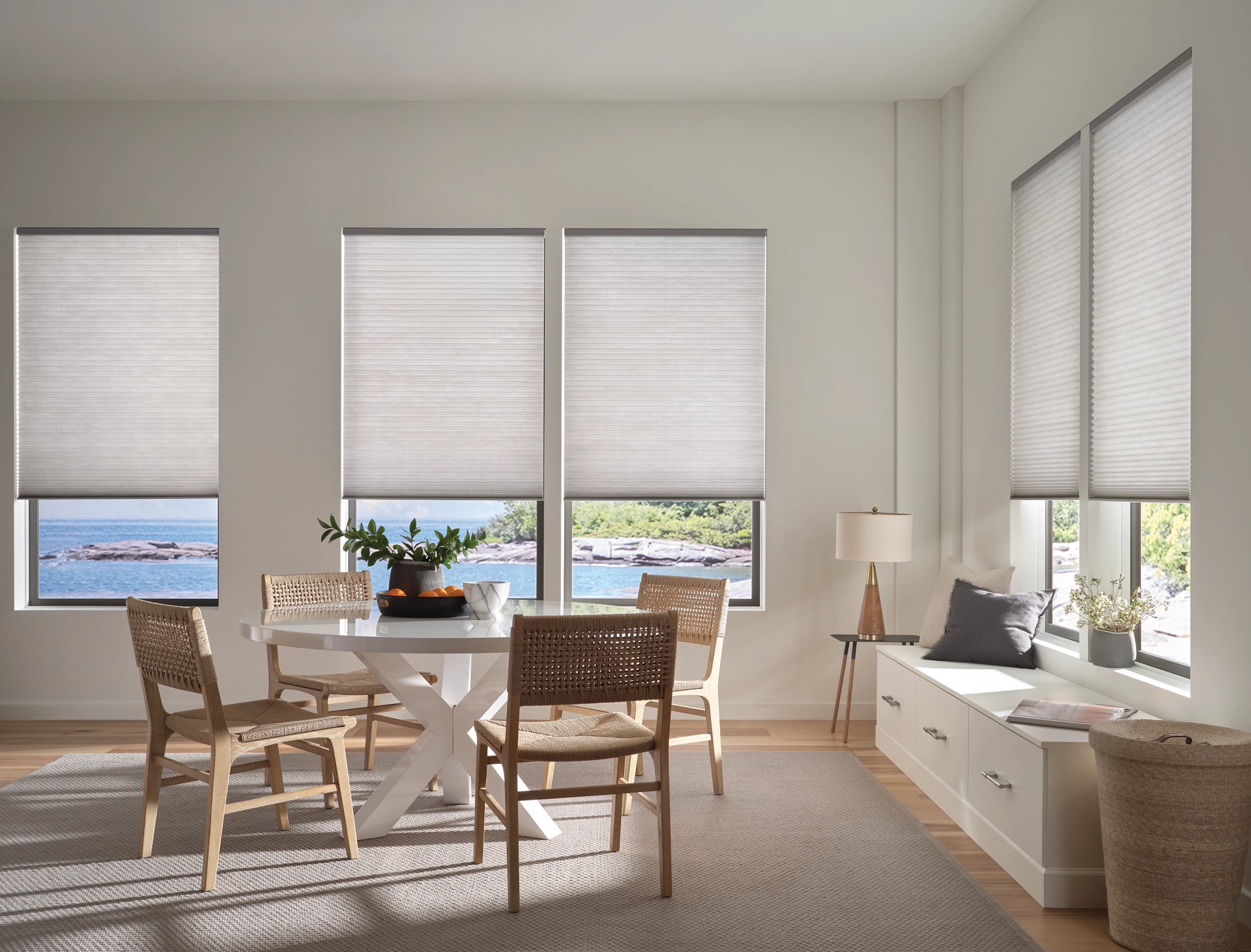 Bright living room with dining table, white blinds, and a bench by large windows overlooking water.