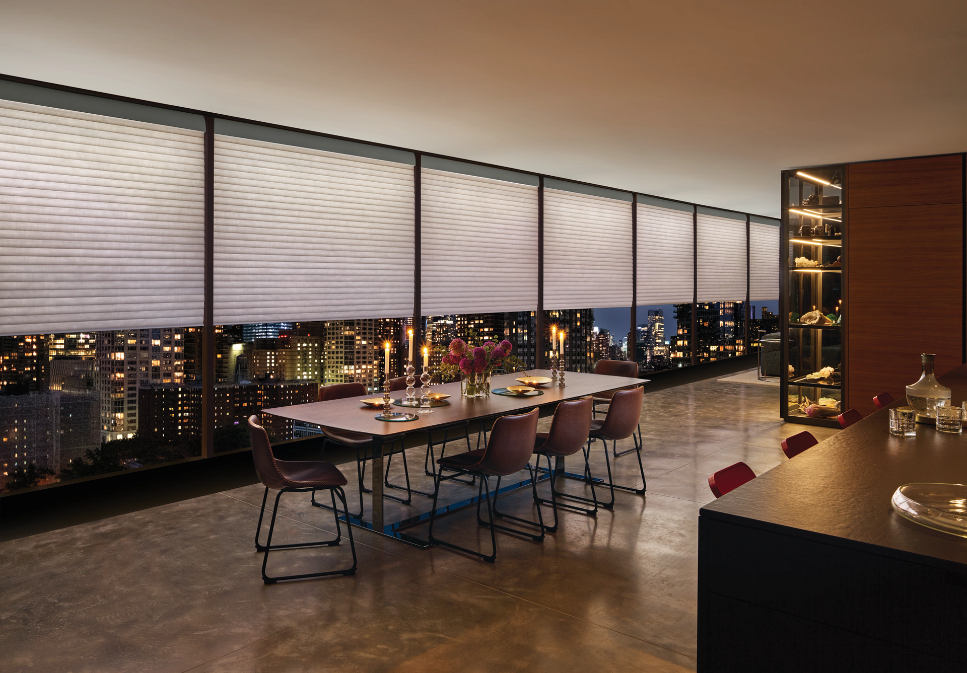 Rooftop dining area with a long table, city lights beyond floor-to-ceiling windows at dusk