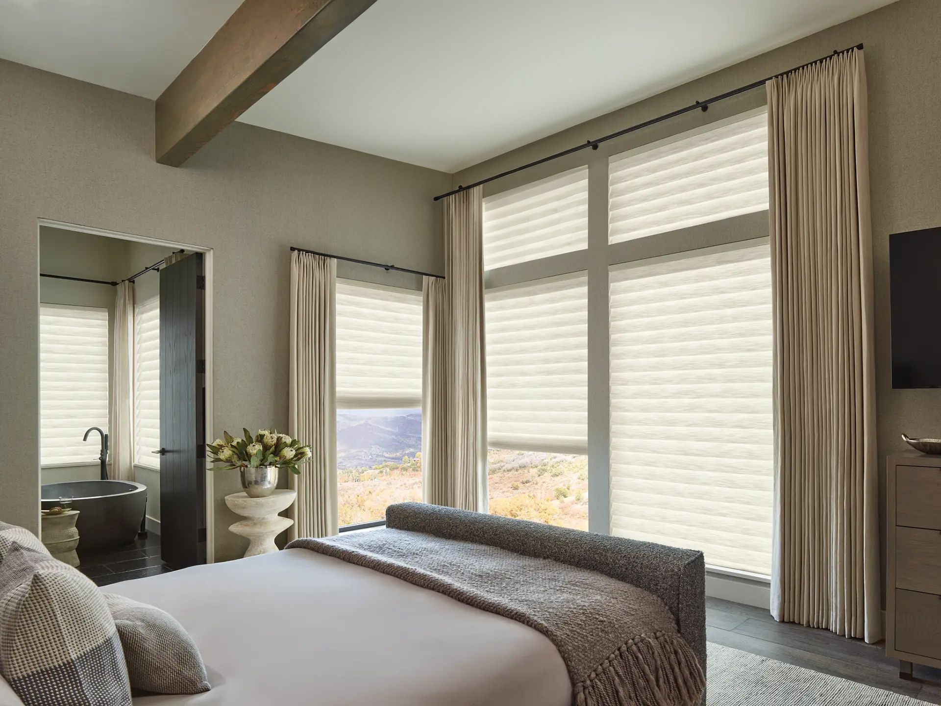 Bright bedroom with large windows, beige curtains, bed, mirror, and a TV on the wall