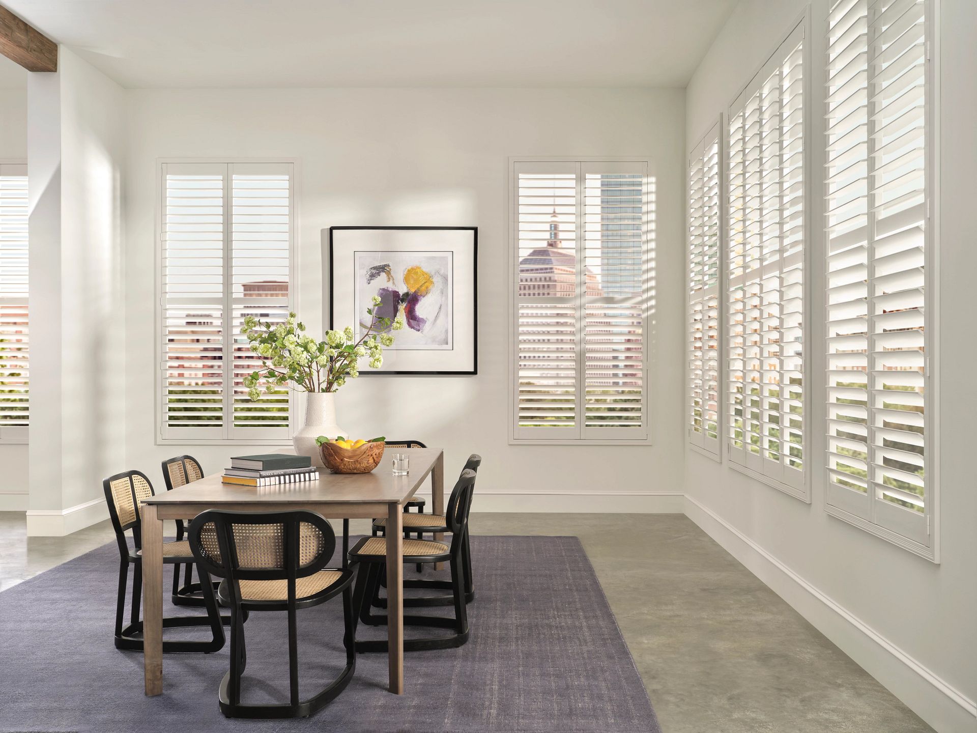 Bright dining room with a wooden table, black chairs, a vase of flowers, and large white shuttered windows