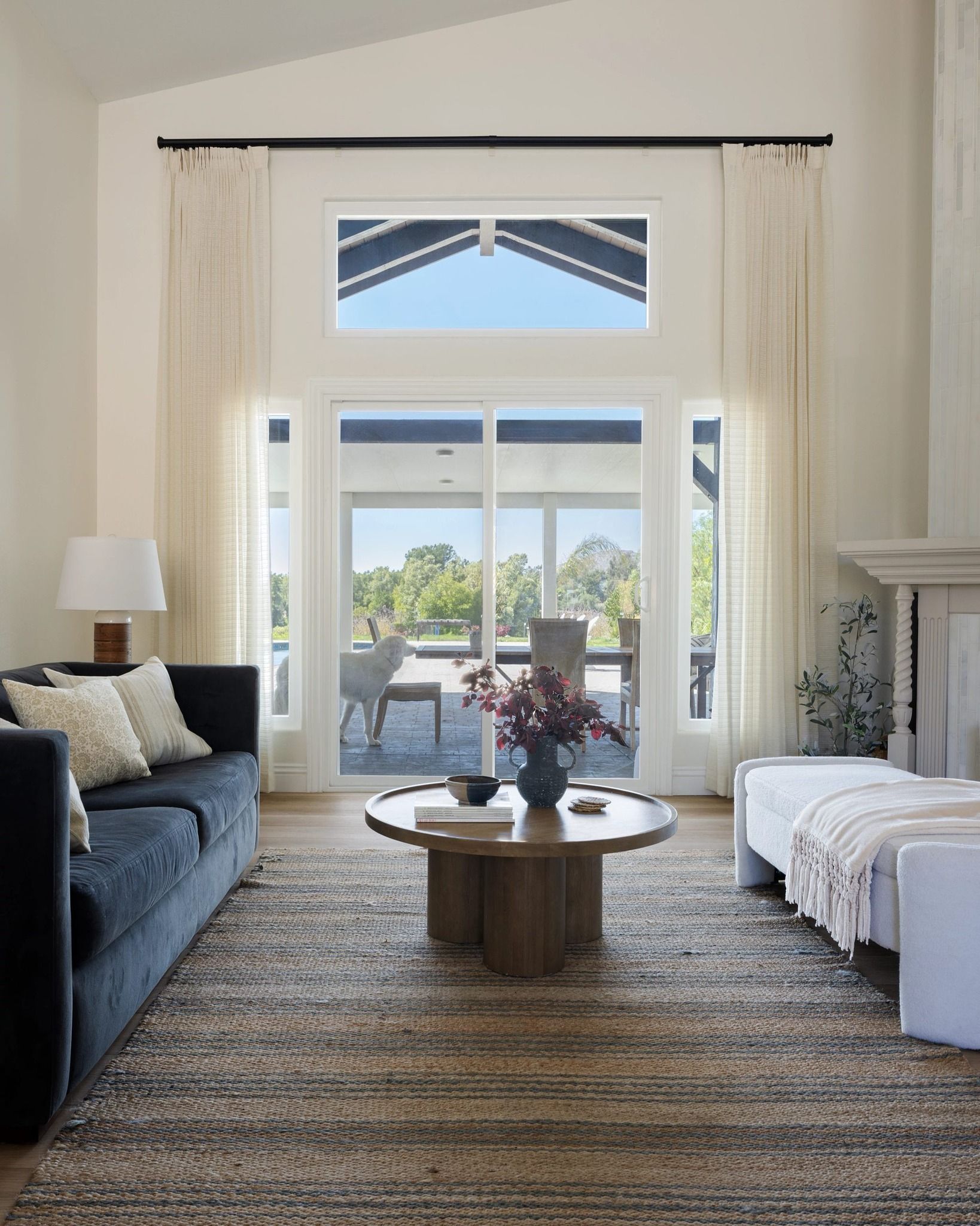 Bright living room with a dark sofa, round coffee table, white bench, and tall windows to a patio.