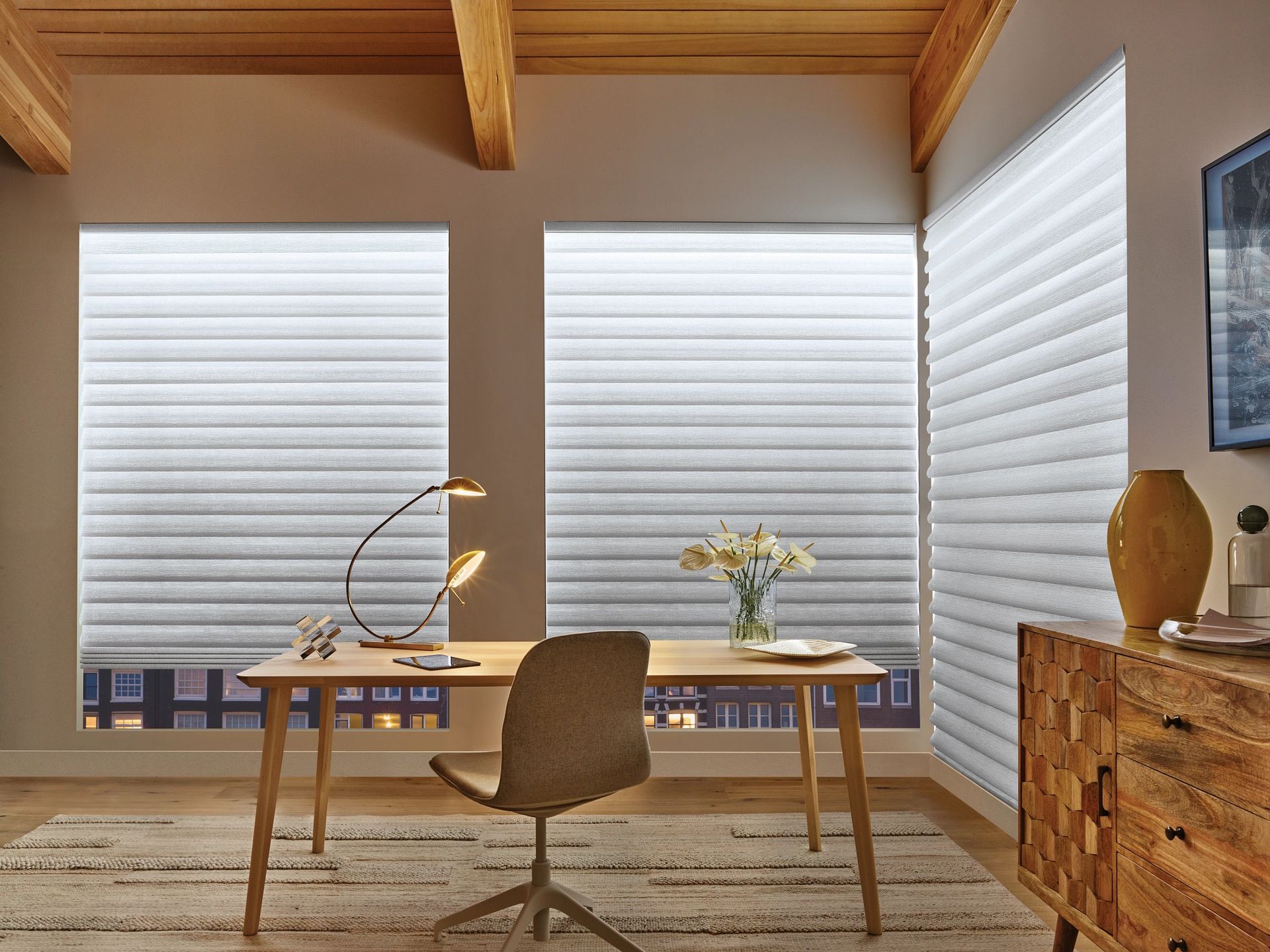 Modern home office with wooden desk, chair, lamp, flowers, and large white blinds.