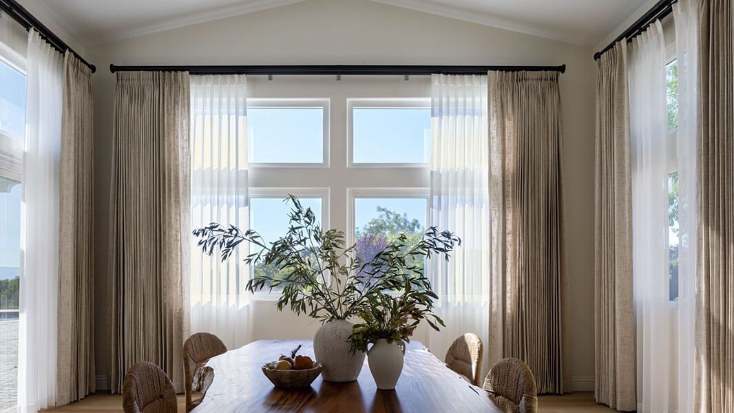 Bright dining room with a wooden table, potted plants, and sheer curtains around large windows.