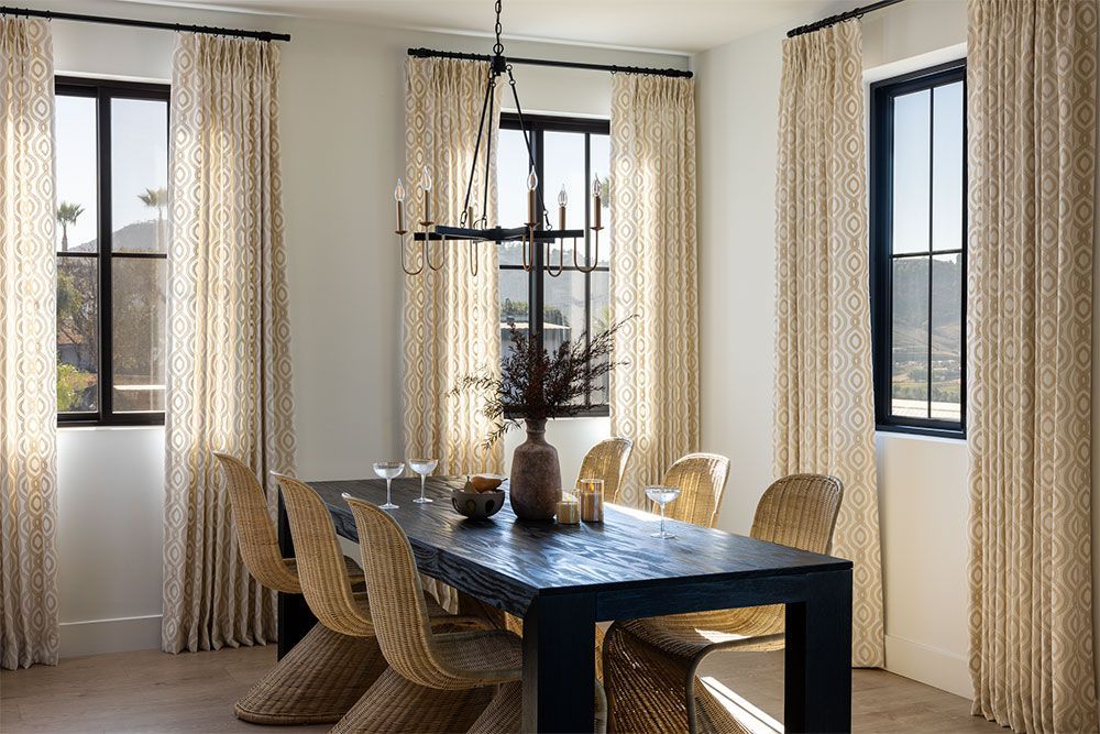 Dining room with a dark table, woven chairs, and beige curtains beside large windows.