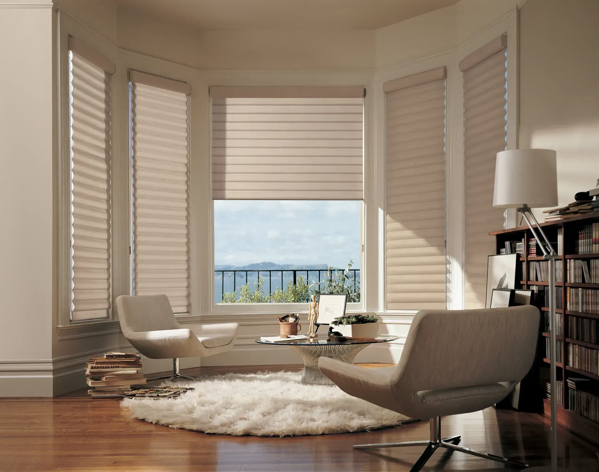 Bright sunlit living room with large windows, white chairs, bookshelf, and a view of the water.