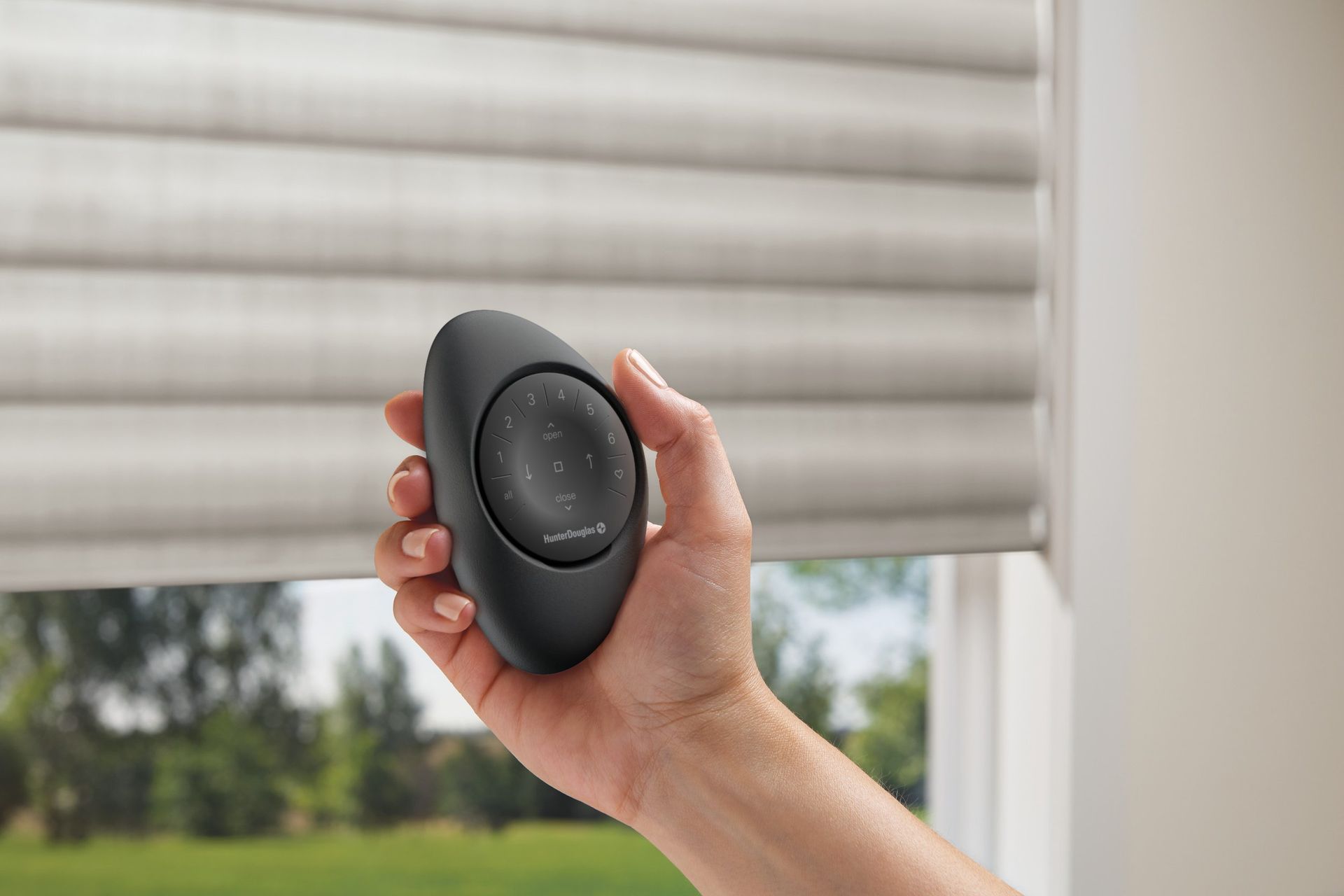 Hand holding a black remote control beside white window blinds with greenery outside