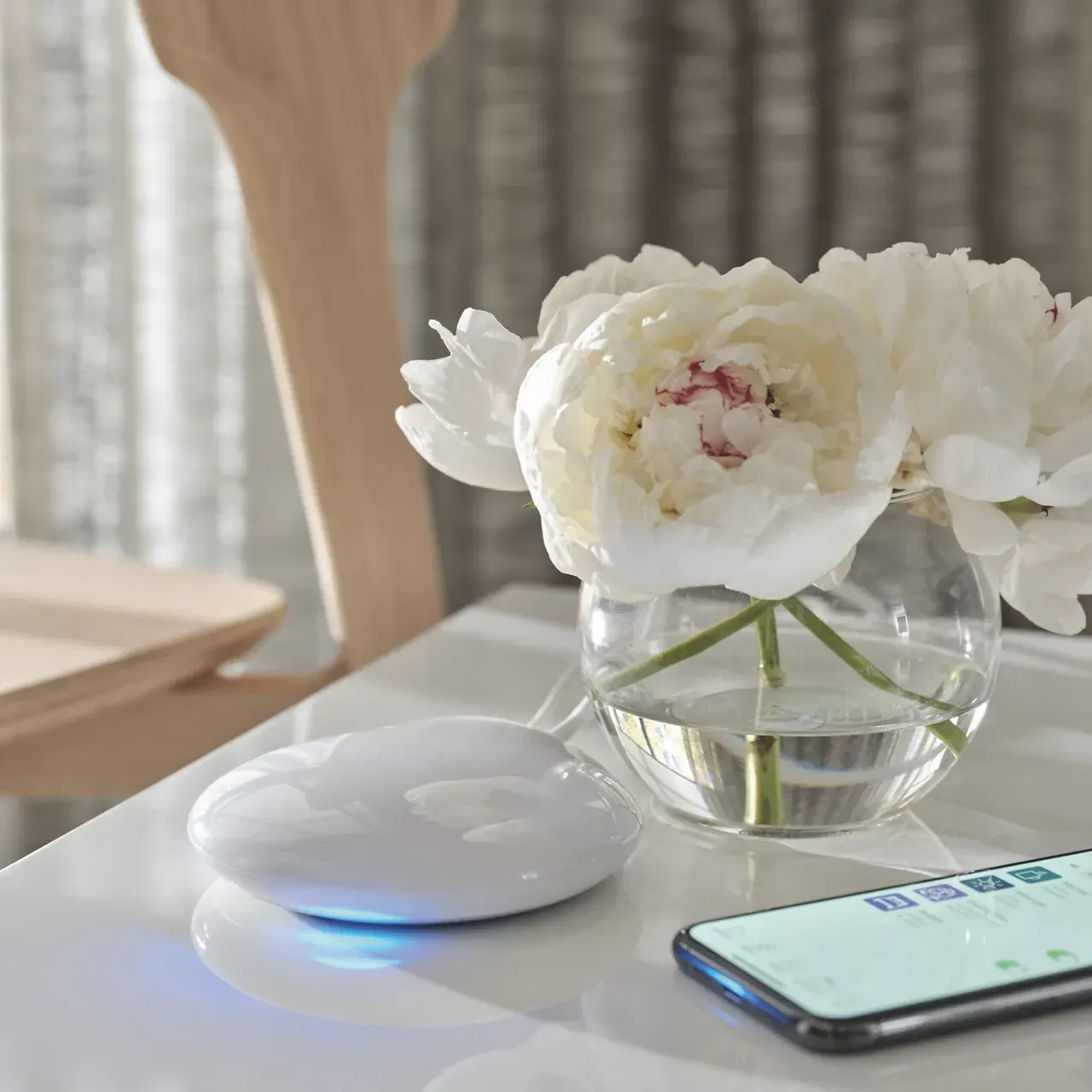 White flowers in a glass vase beside a white device and smartphone on a sunlit table