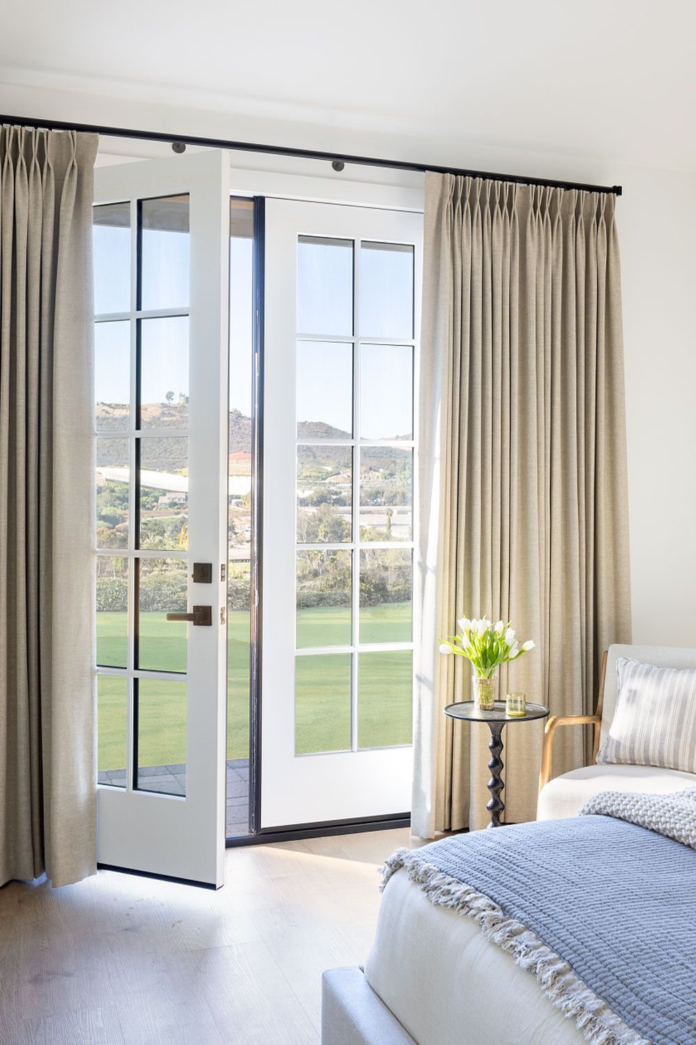 Bright bedroom with open French doors, beige curtains, and a bed by a sunlit window overlooking green hills
