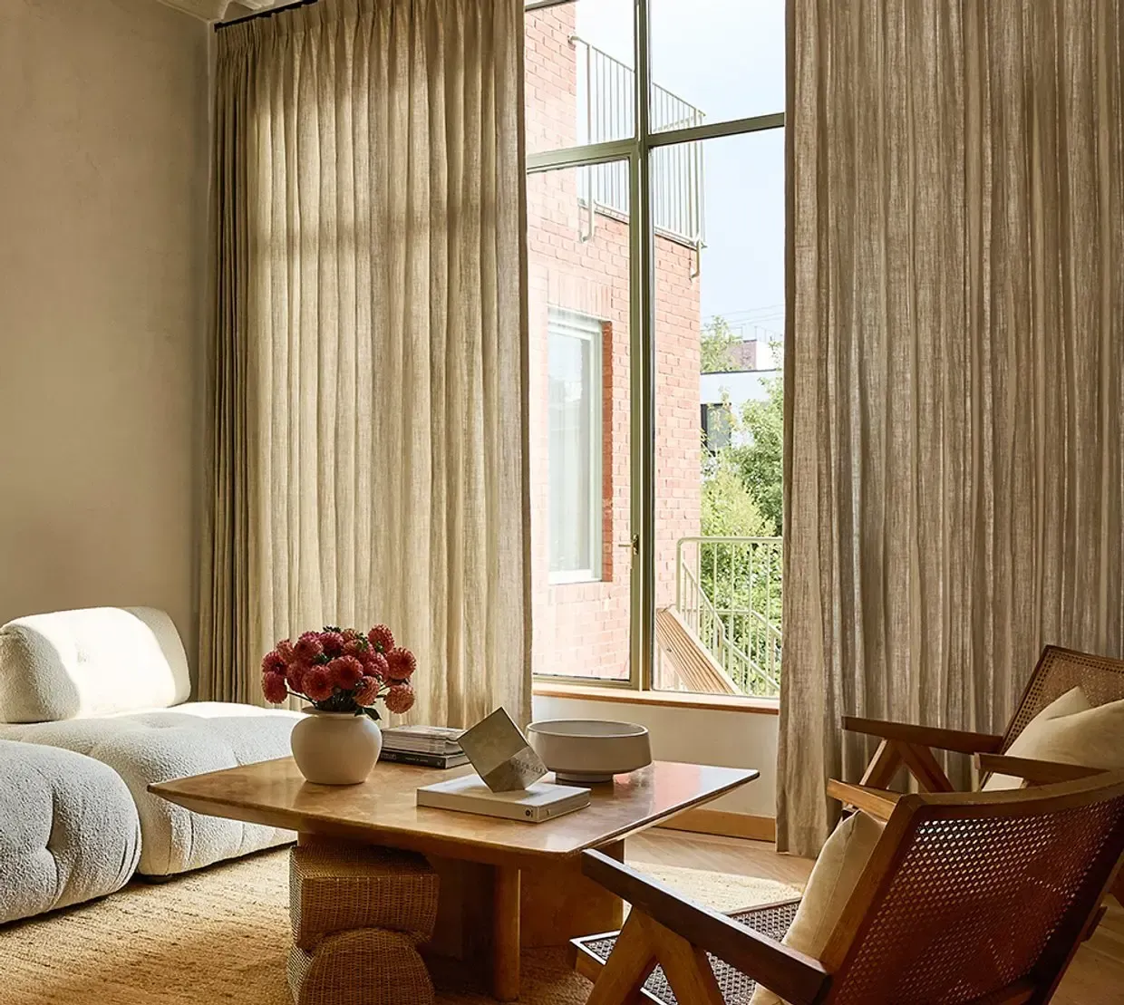 Sunlit living room with beige curtains, wooden chairs, and a coffee table by open balcony doors