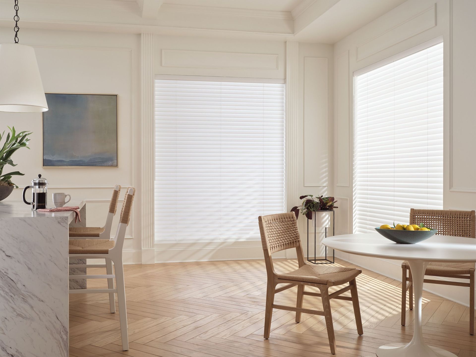 Bright dining room with wicker chairs, white table, and sunlight through large windows with blinds