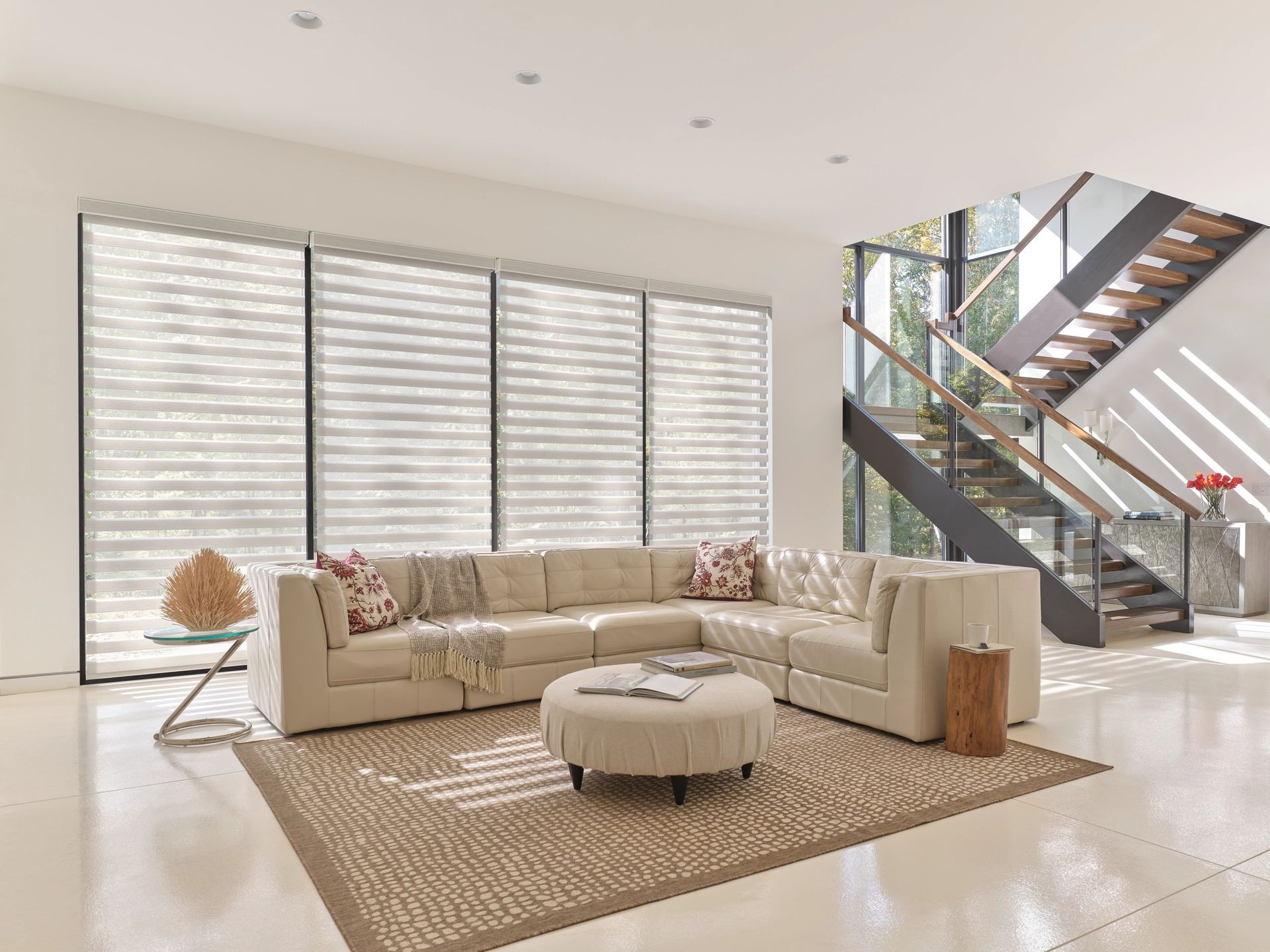 Modern living room with beige sectional sofa, round ottoman, woven rug, and staircase with glass railing