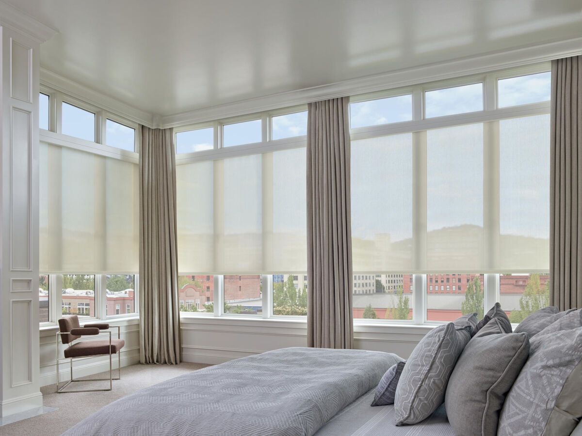 Bright bedroom with floor-to-ceiling windows, gray bedding, and beige curtains overlooking a city view