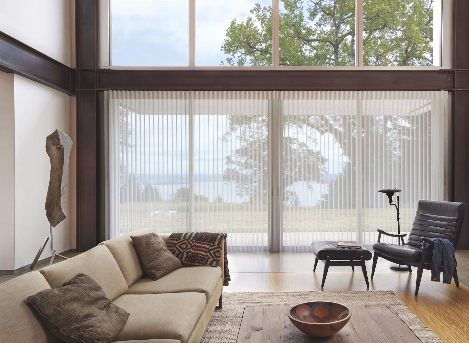 Modern living room with beige sofa, tall windows, sheer curtains, and dark chairs around a wooden floor