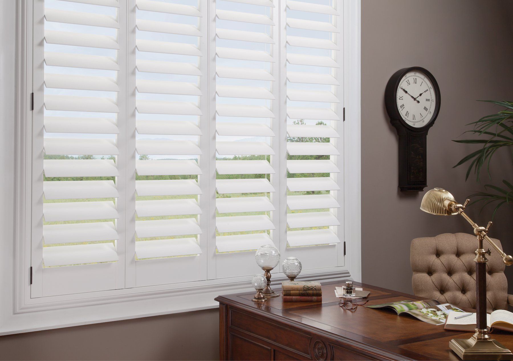 Home office with white shutters, wooden desk, tufted chair, wall clock, and lamp