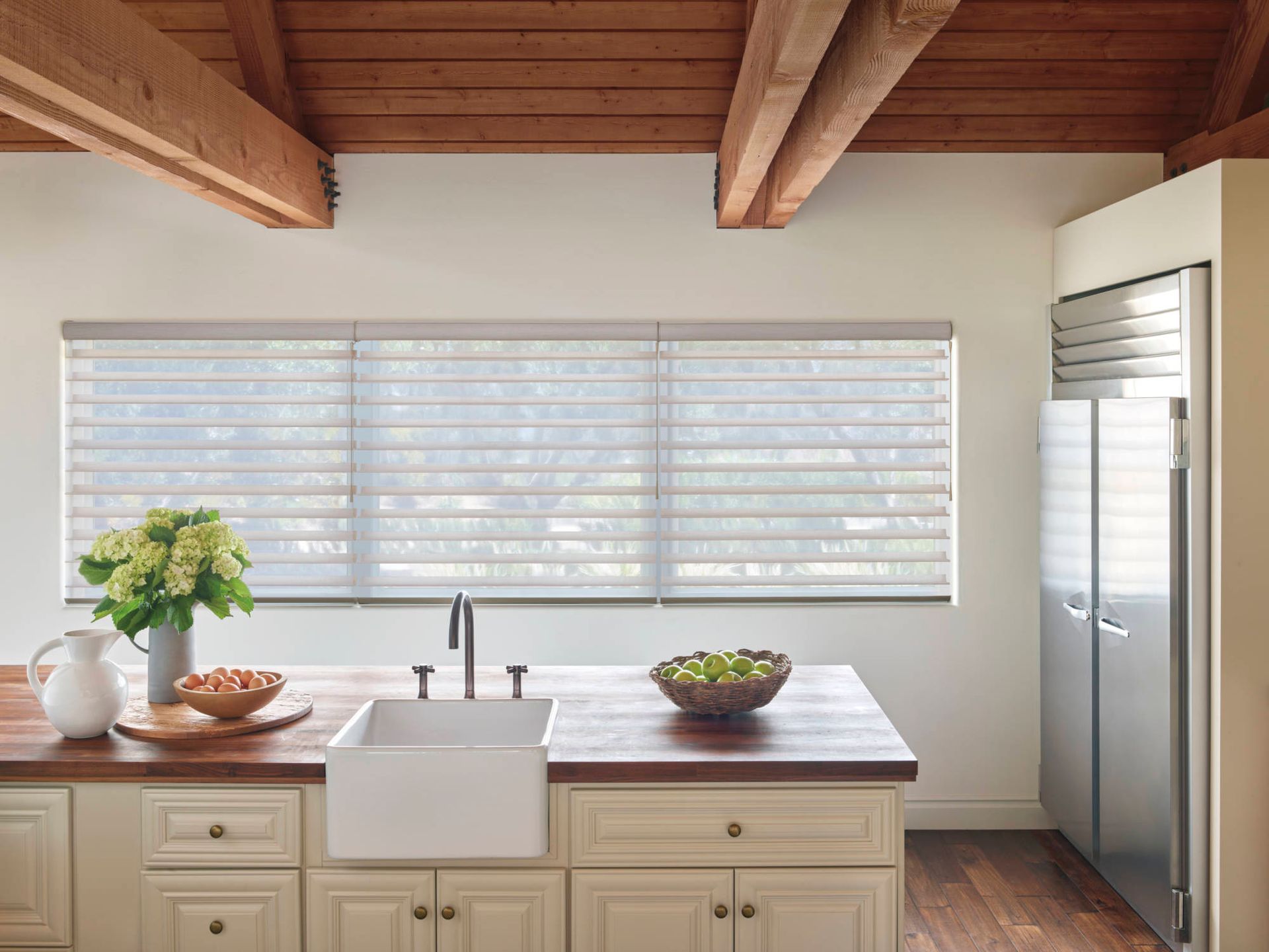 Bright kitchen with white cabinets, farmhouse sink, large window blinds, and stainless steel refrigerator.