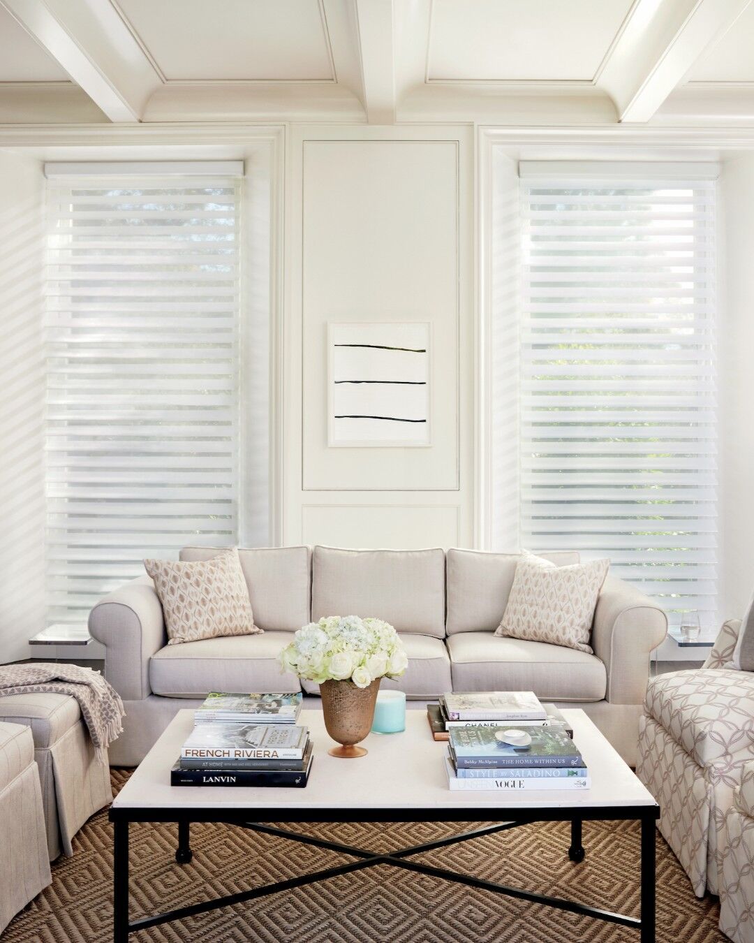 Bright living room with a beige sofa, two armchairs, white coffee table, and tall windows with blinds.