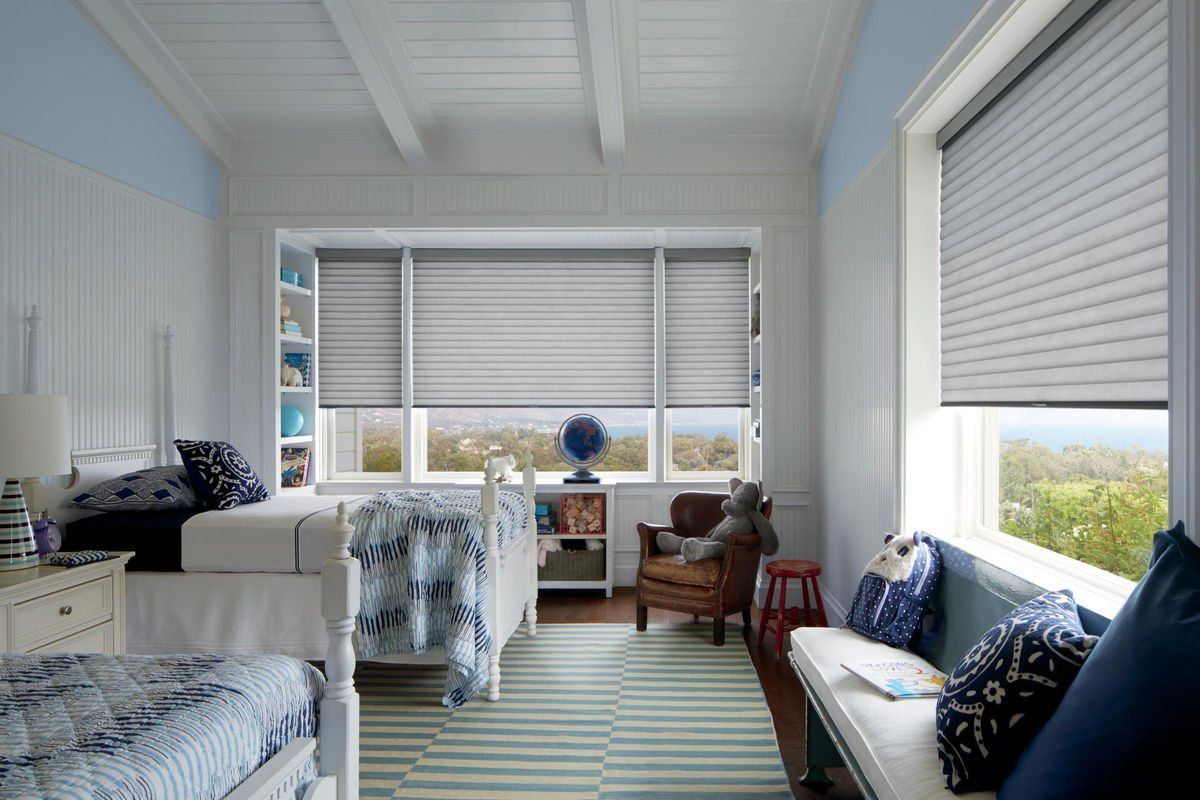 Bright sunroom with twin beds, blue accents, striped rug, and white blinds overlooking greenery.