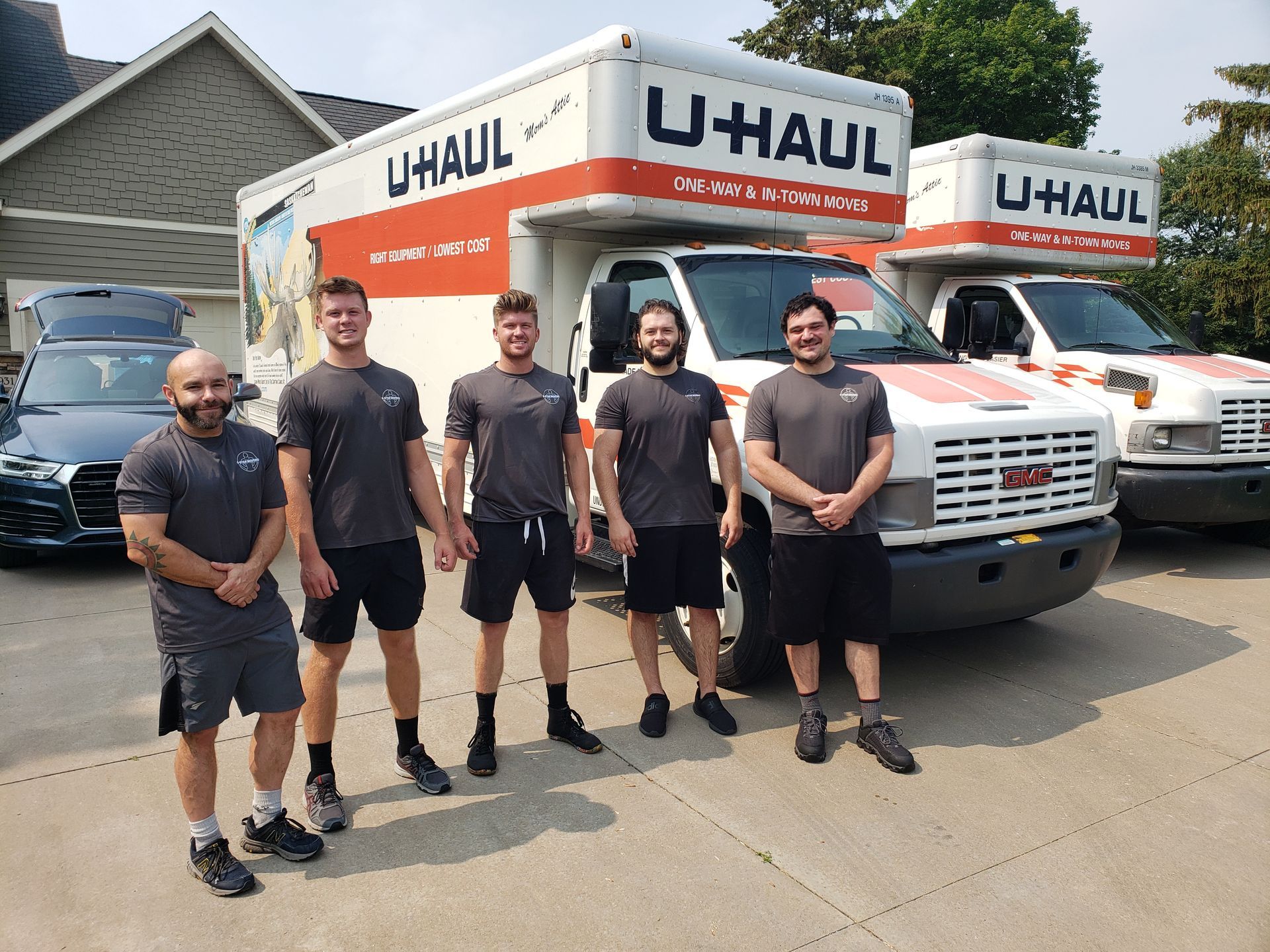 Five men stand in front of U-Haul trucks on a sunny day. They wear gray shirts and black shorts.