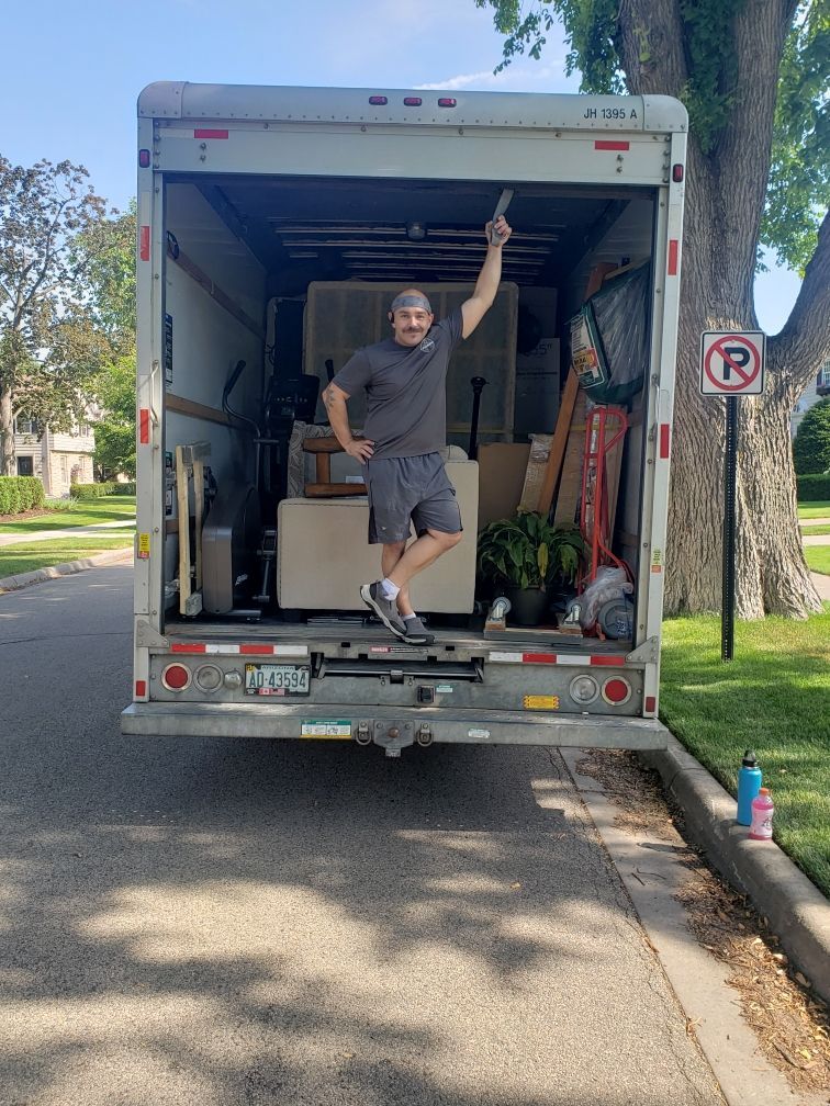 Man stands in back of moving truck, arms raised in triumph on sunny street.