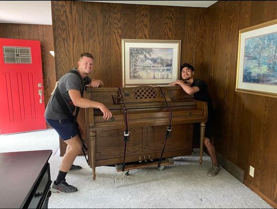 Two people moving an upright wooden piano on a dolly in a room with wood-paneled walls and a red door.