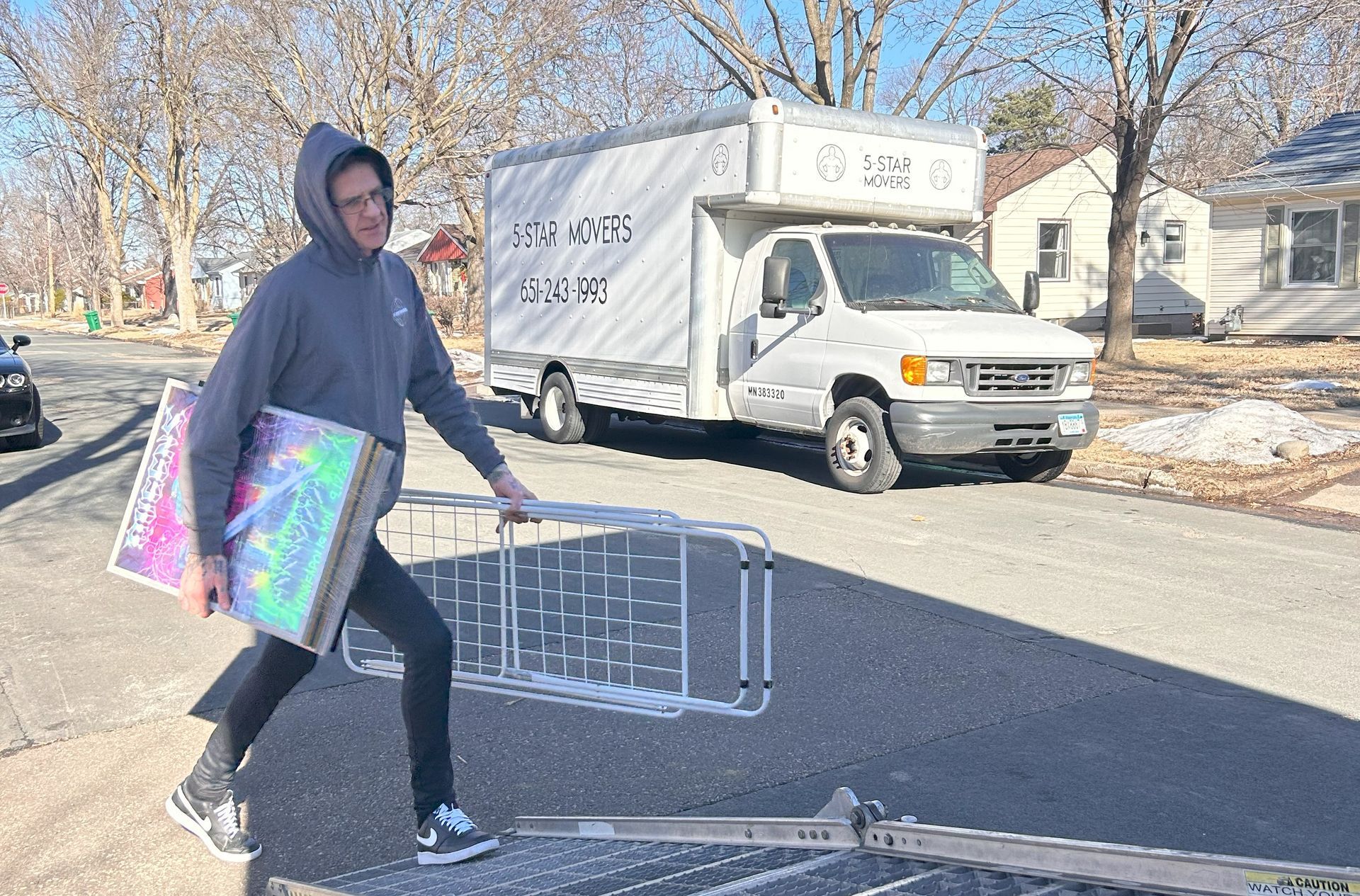 Man carrying items from a moving truck parked on a residential street. Sunlight, gray truck, and snow present.