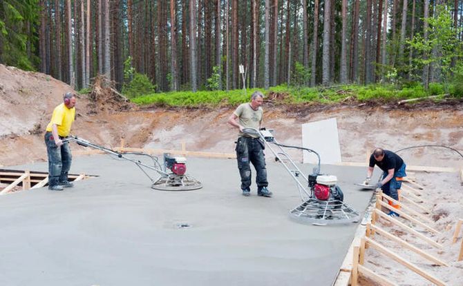 Three workers smoothing wet concrete at a forested construction site with power trowels