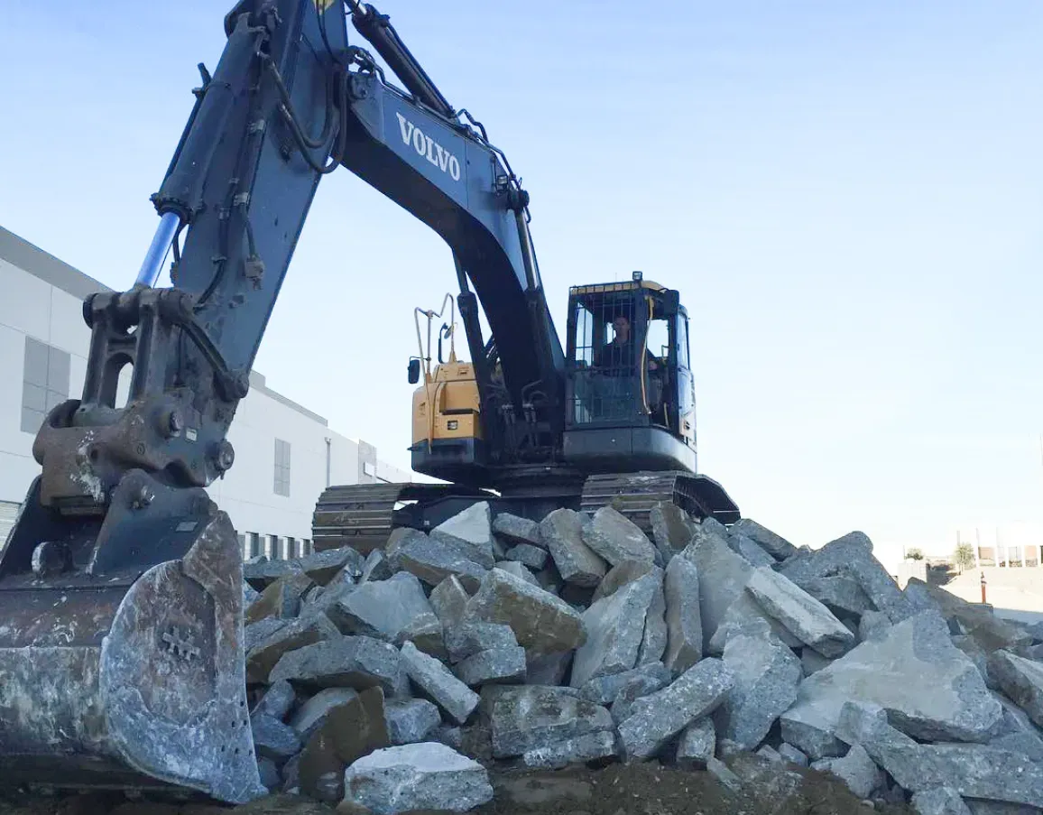 A dark blue Volvo excavator positioned on top of a large pile of broken concrete and stone at a construction site.