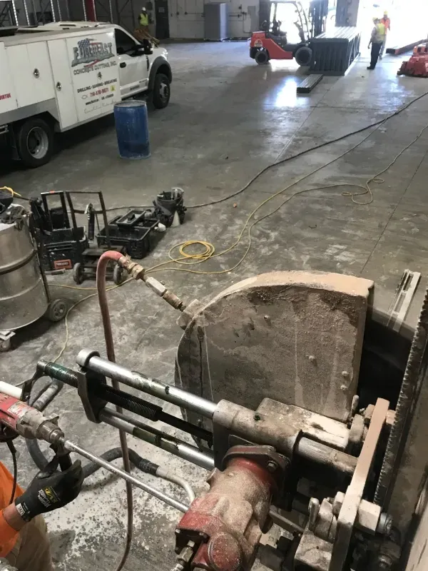 A person operates a handheld concrete saw in a warehouse with a utility truck and construction equipment in the background.