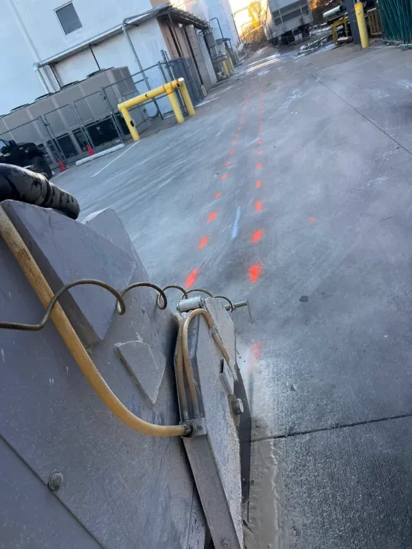 A concrete saw operates on a parking lot, following a line of orange spray-painted marks toward a building.