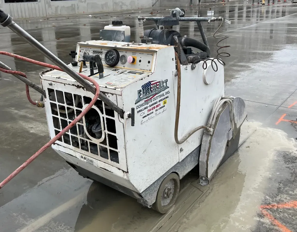 A white walk-behind electric concrete flat saw sits on a wet, paved construction site, cutting a groove in the surface.