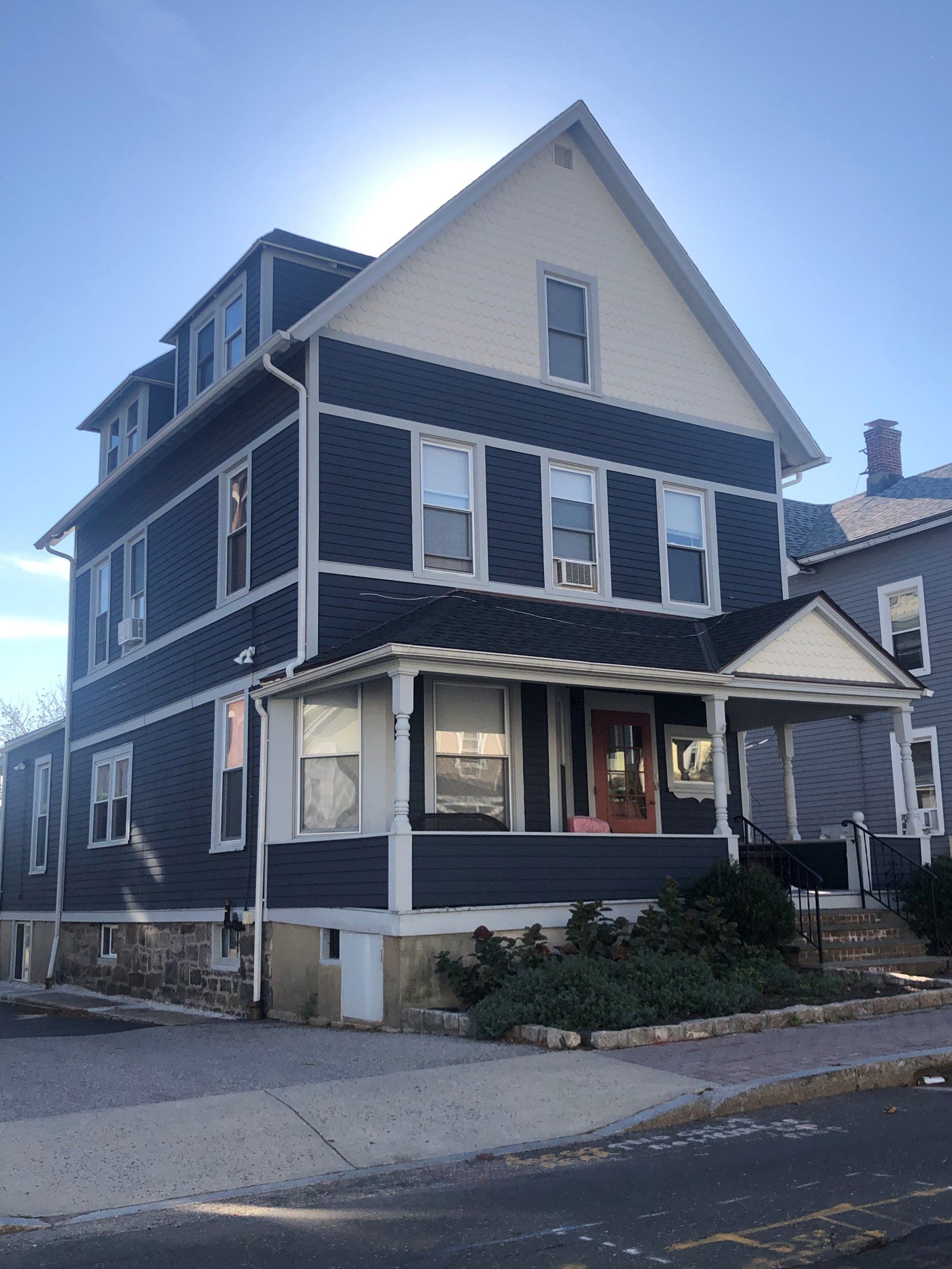 A three-story, blue-sided house with a white trim, front porch, and a dormer window under a sunny sky.